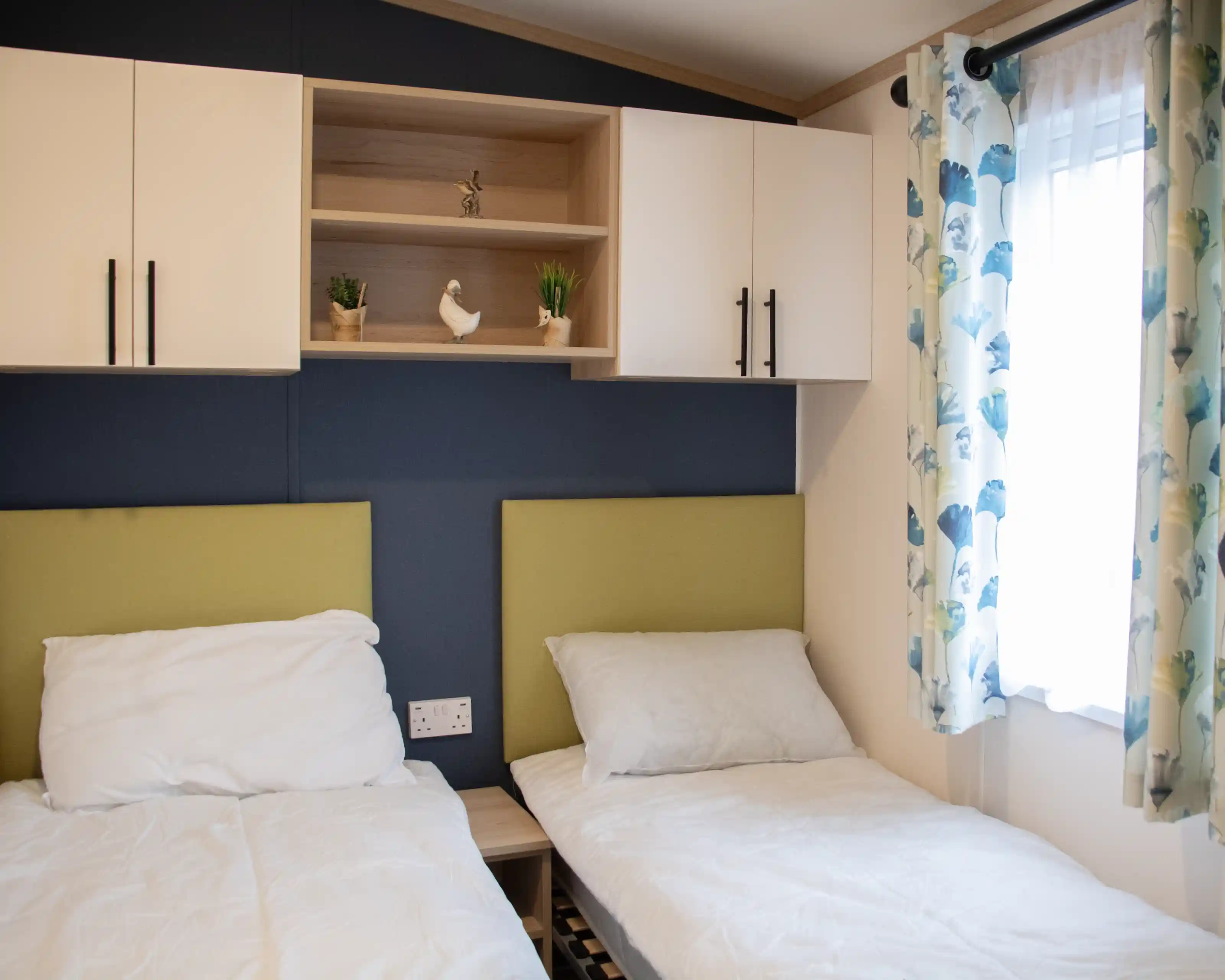 Two single beds with white bedding are positioned against a blue wall. Above the beds, a wooden shelf holds decorative plants and a small ornamental bird. A window with light curtains adorned with a floral pattern adds natural light to the room.