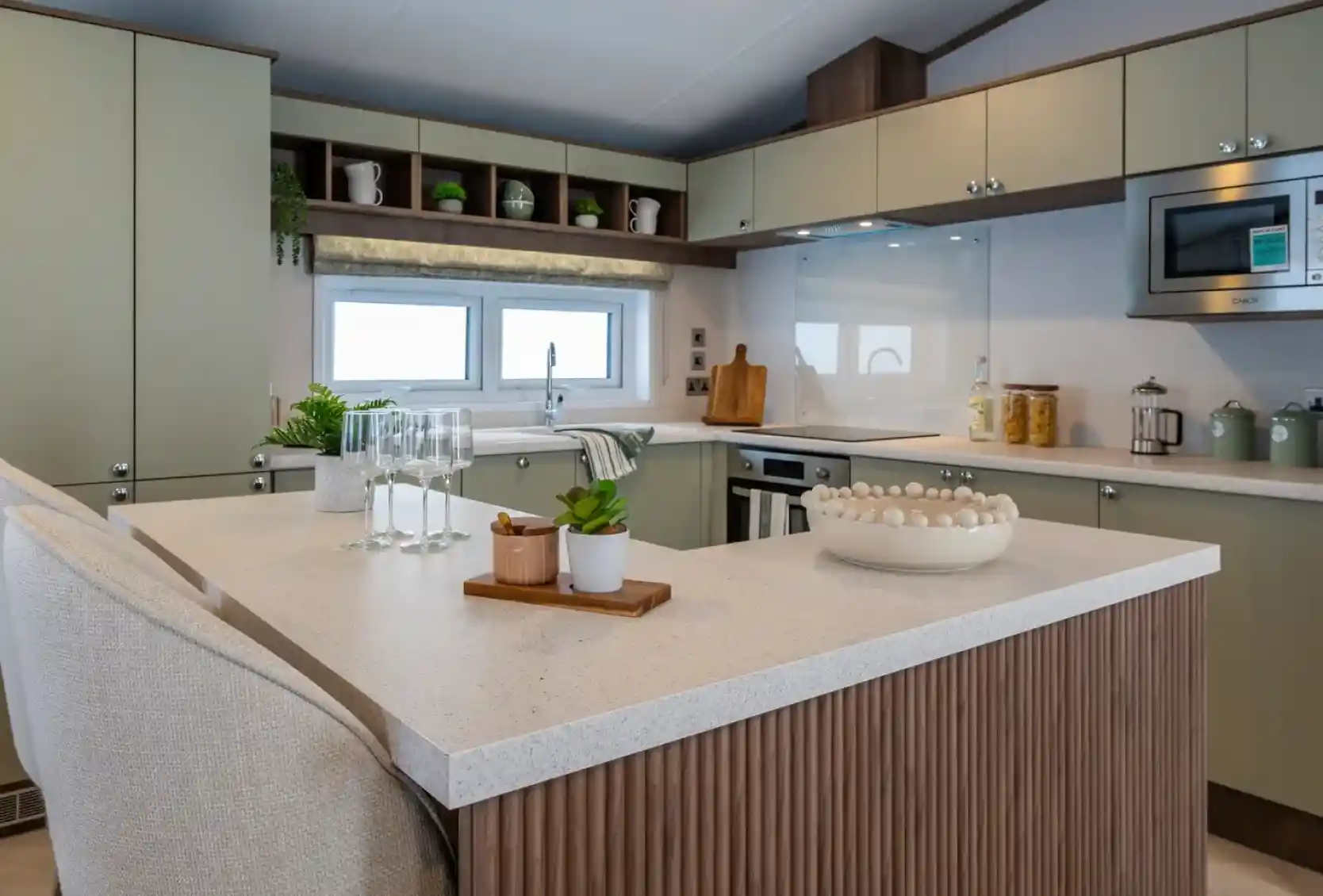 Modern kitchen featuring green cabinets, a marble countertop, and a central island. Glassware and a small potted plant are on the island, along with a decorative bowl. Shelves above display kitchenware, with a window providing natural light.