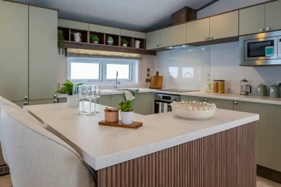 Modern kitchen featuring green cabinets, a marble countertop, and a central island. Glassware and a small potted plant are on the island, along with a decorative bowl. Shelves above display kitchenware, with a window providing natural light.