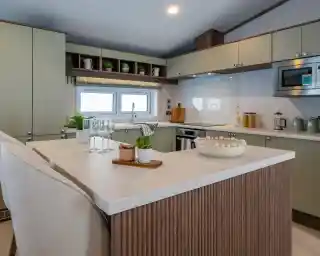 Modern kitchen featuring green cabinets, a marble countertop, and a central island. Glassware and a small potted plant are on the island, along with a decorative bowl. Shelves above display kitchenware, with a window providing natural light.