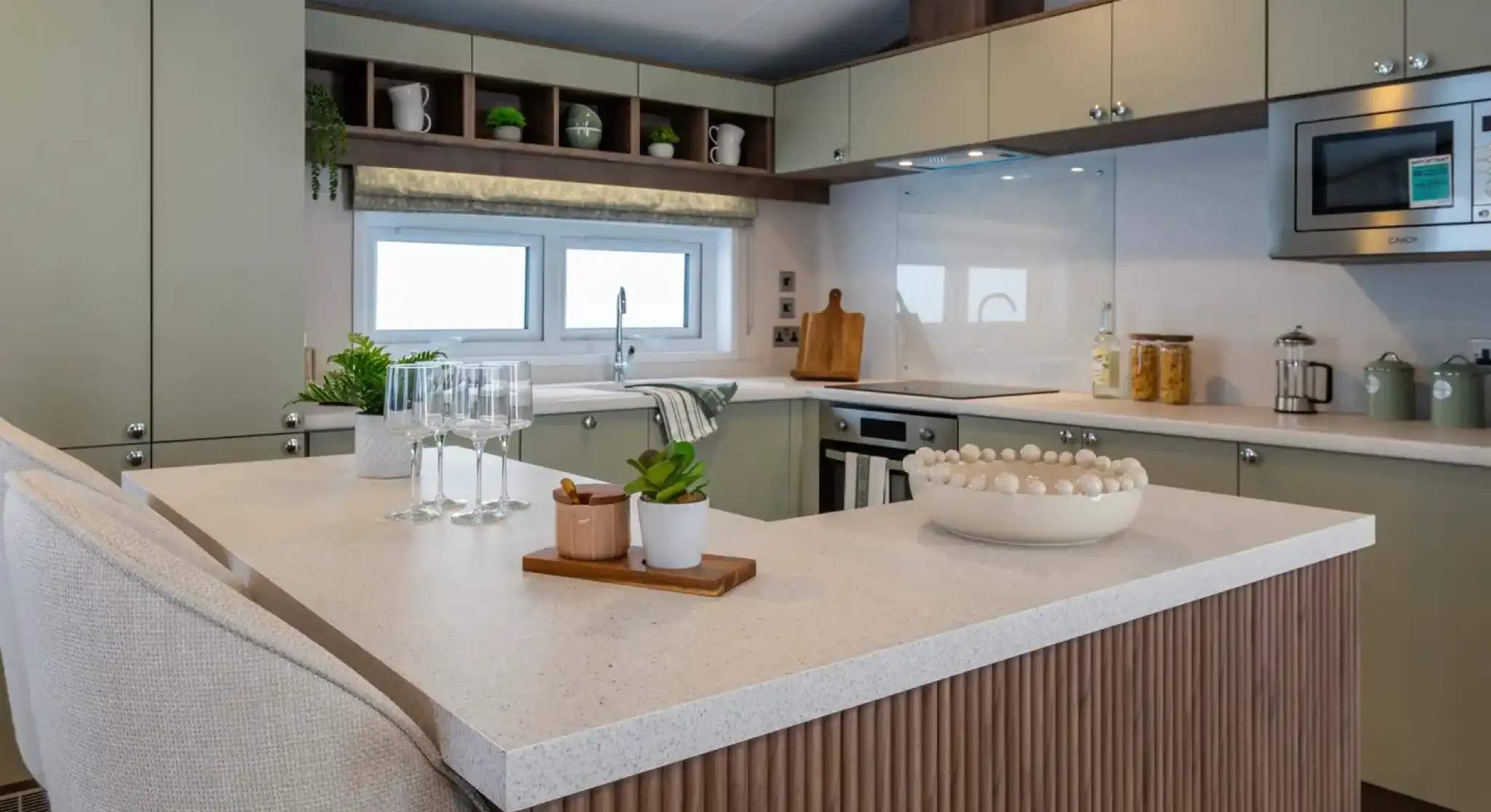 Modern kitchen featuring green cabinets, a marble countertop, and a central island. Glassware and a small potted plant are on the island, along with a decorative bowl. Shelves above display kitchenware, with a window providing natural light.