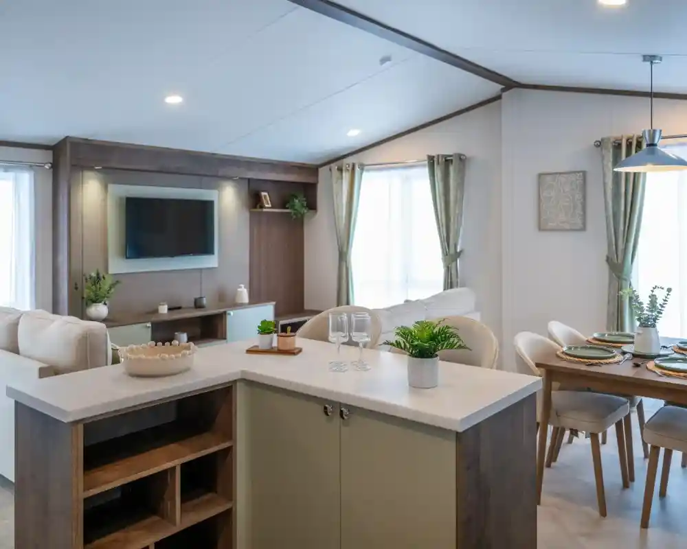 A modern interior of a living space featuring a cozy sofa, a mounted television, and a dining area with a wooden table set for four. Natural light fills the room through large windows, and decorative plants add a touch of greenery.