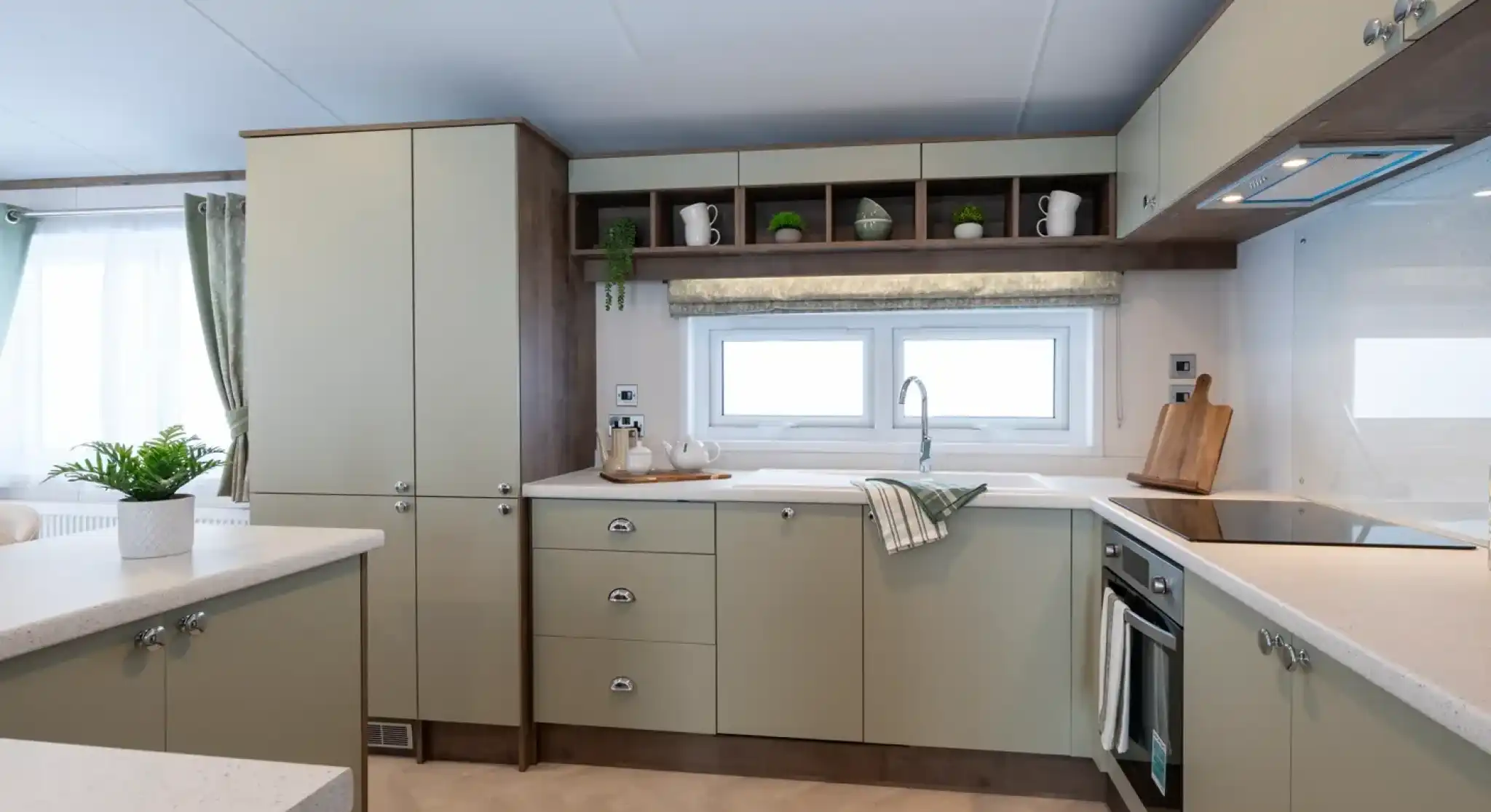 Modern kitchen with light green cabinets, white countertops, and stainless steel appliances. A window lets in natural light, and decorative items are arranged on the shelves.