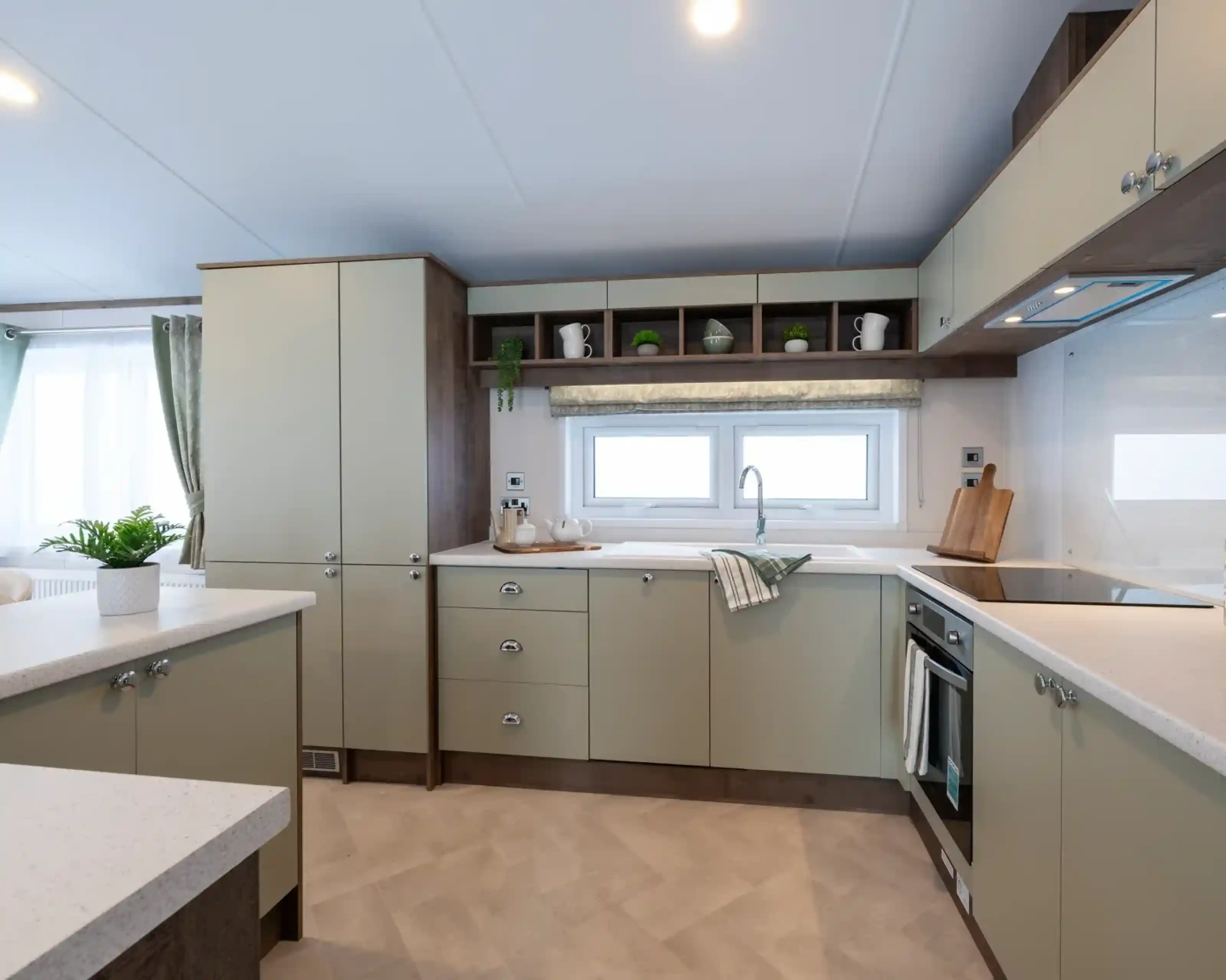 Modern kitchen with light green cabinets, white countertops, and stainless steel appliances. A window lets in natural light, and decorative items are arranged on the shelves.