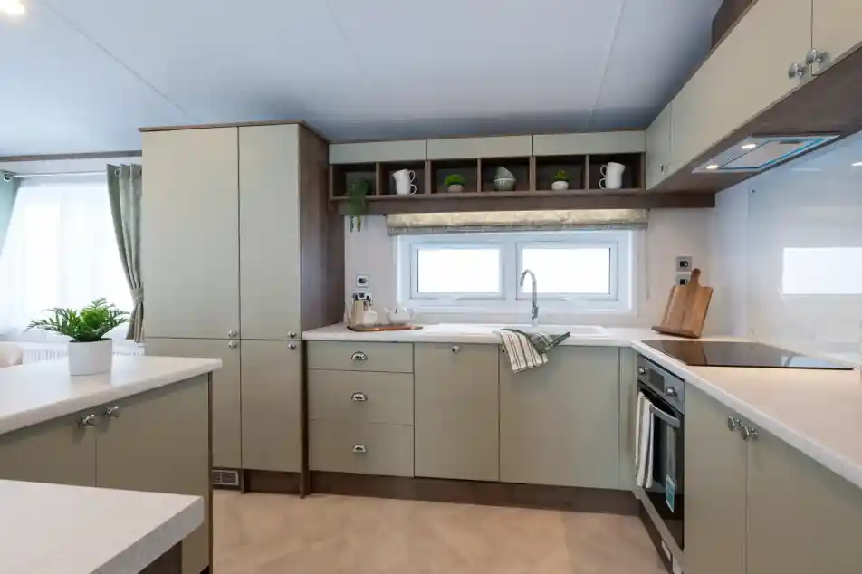 Modern kitchen with light green cabinets, white countertops, and stainless steel appliances. A window lets in natural light, and decorative items are arranged on the shelves.