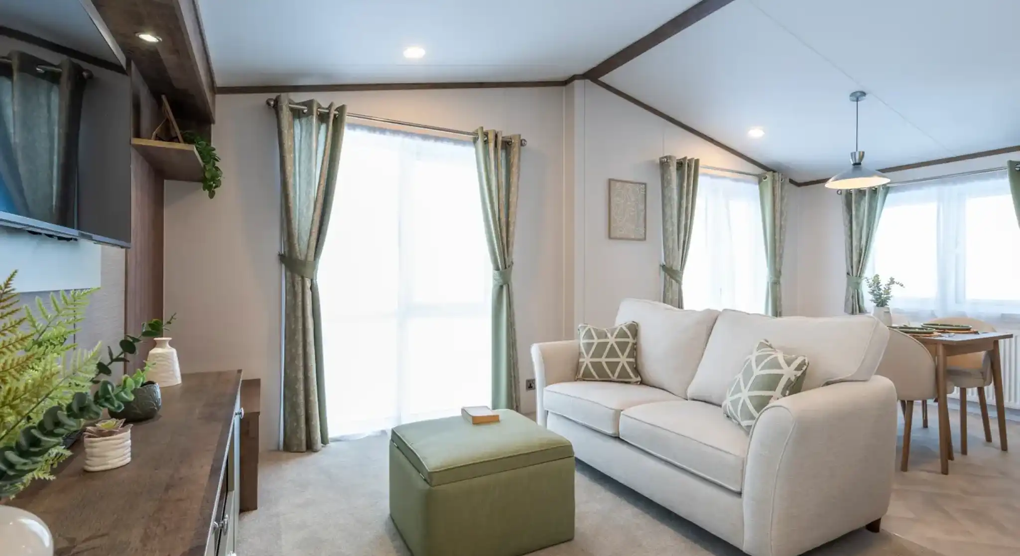 A cozy living room featuring a light-colored sofa with decorative pillows, an olive green ottoman, and large windows dressed in sheer curtains. A small dining area with a table and chairs is visible in the background, and a dark wooden entertainment unit holds decorative items. The room is bright and welcoming with a soft carpet.