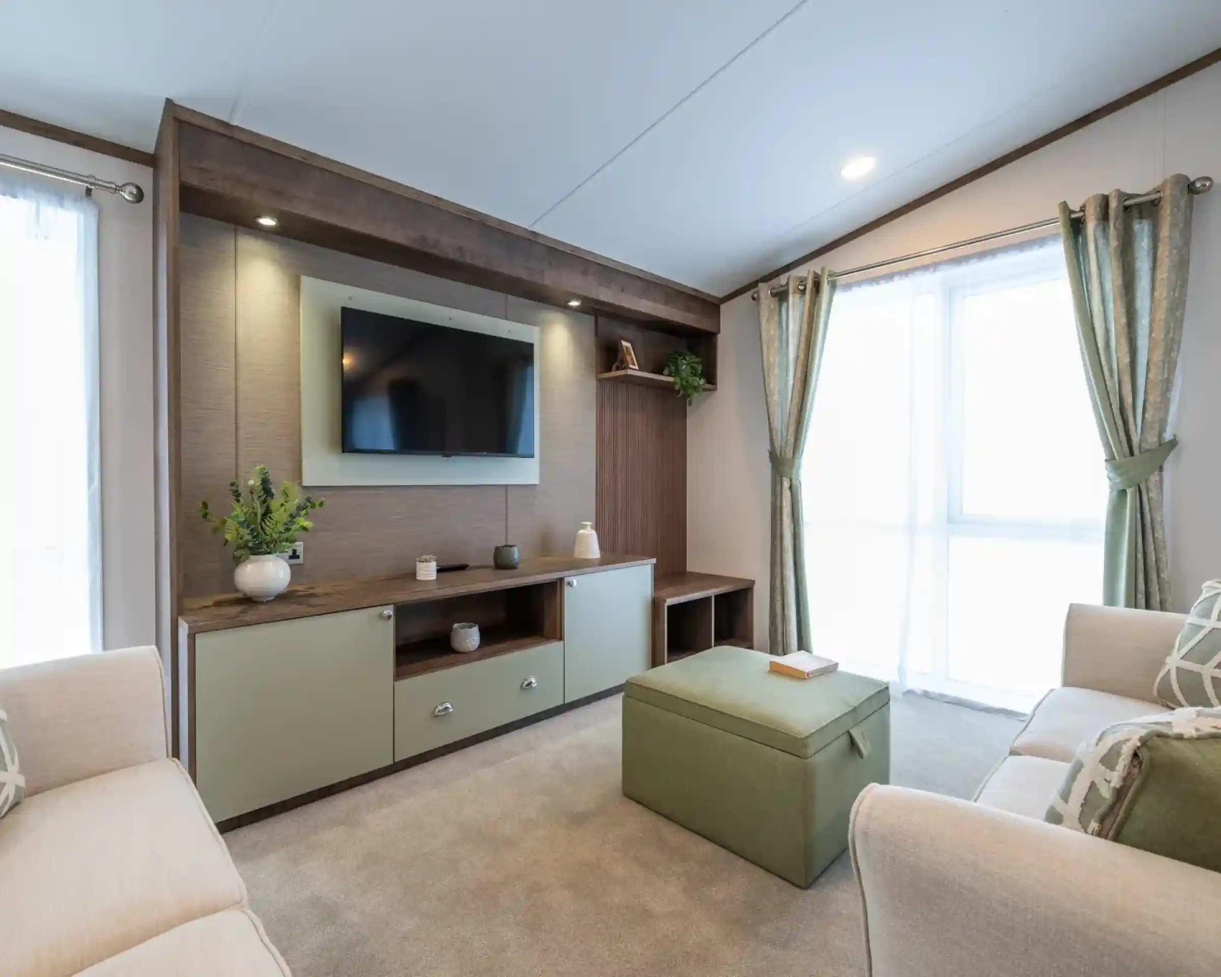 A modern living room featuring a wall-mounted TV above a wooden console with decorative items. Light green ottoman in the center and two beige sofas flanking either side. Large windows with sheer curtains allow natural light to fill the space.