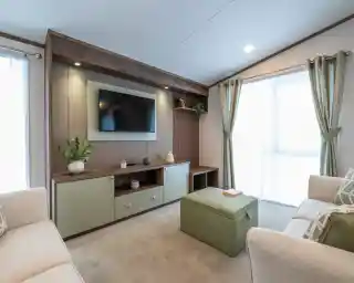 A modern living room featuring a wall-mounted TV above a wooden console with decorative items. Light green ottoman in the center and two beige sofas flanking either side. Large windows with sheer curtains allow natural light to fill the space.