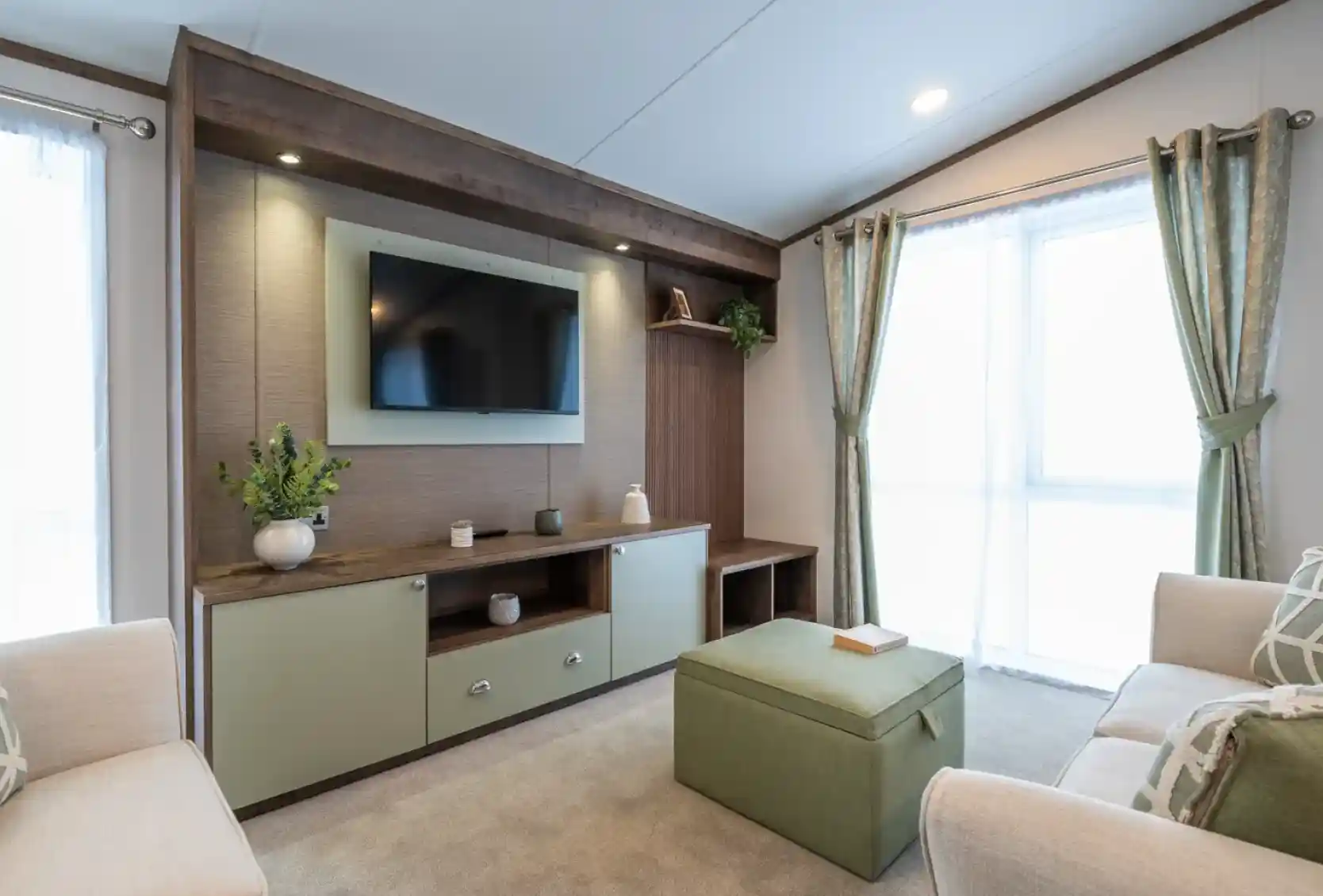 A modern living room featuring a wall-mounted TV above a wooden console with decorative items. Light green ottoman in the center and two beige sofas flanking either side. Large windows with sheer curtains allow natural light to fill the space.