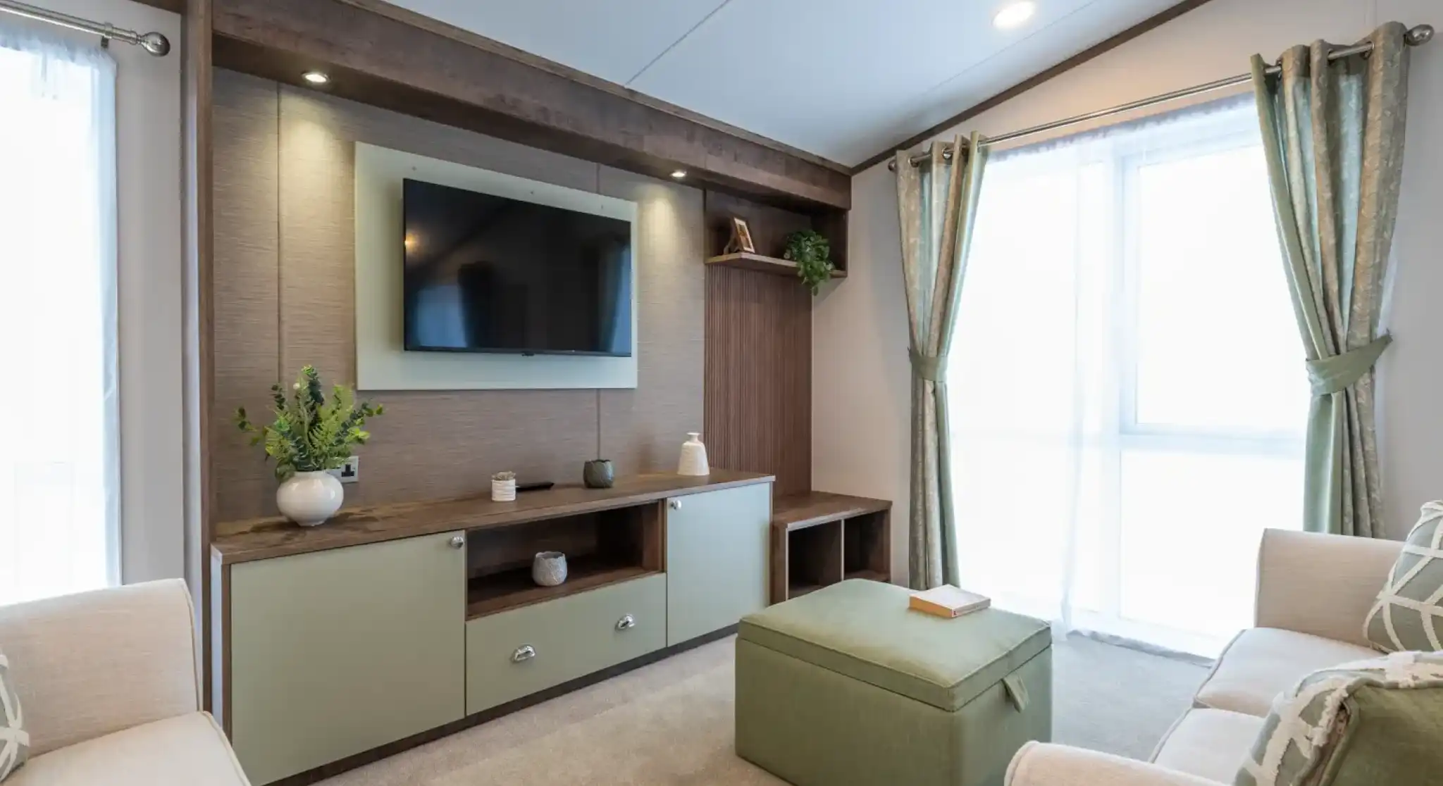A modern living room featuring a wall-mounted TV above a wooden console with decorative items. Light green ottoman in the center and two beige sofas flanking either side. Large windows with sheer curtains allow natural light to fill the space.