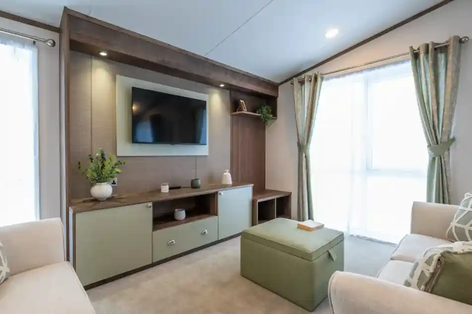 A modern living room featuring a wall-mounted TV above a wooden console with decorative items. Light green ottoman in the center and two beige sofas flanking either side. Large windows with sheer curtains allow natural light to fill the space.