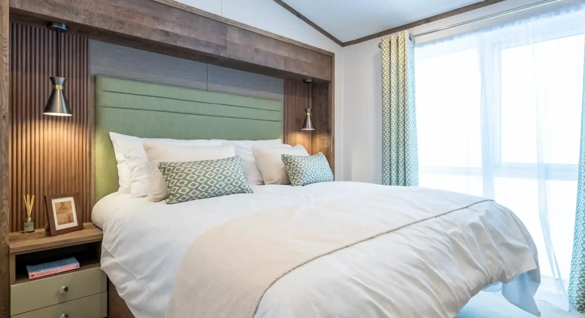A modern bedroom featuring a king-sized bed with a light green headboard and decorative pillows. On either side of the bed are stylish lamps. The room has a neutral color palette with soft lighting and a large window dressed with sheer curtains. A bedside table with decorative items is visible.