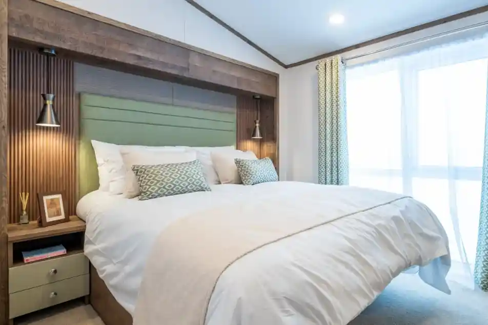 A modern bedroom featuring a king-sized bed with a light green headboard and decorative pillows. On either side of the bed are stylish lamps. The room has a neutral color palette with soft lighting and a large window dressed with sheer curtains. A bedside table with decorative items is visible.