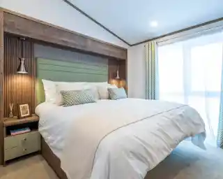 A modern bedroom featuring a king-sized bed with a light green headboard and decorative pillows. On either side of the bed are stylish lamps. The room has a neutral color palette with soft lighting and a large window dressed with sheer curtains. A bedside table with decorative items is visible.