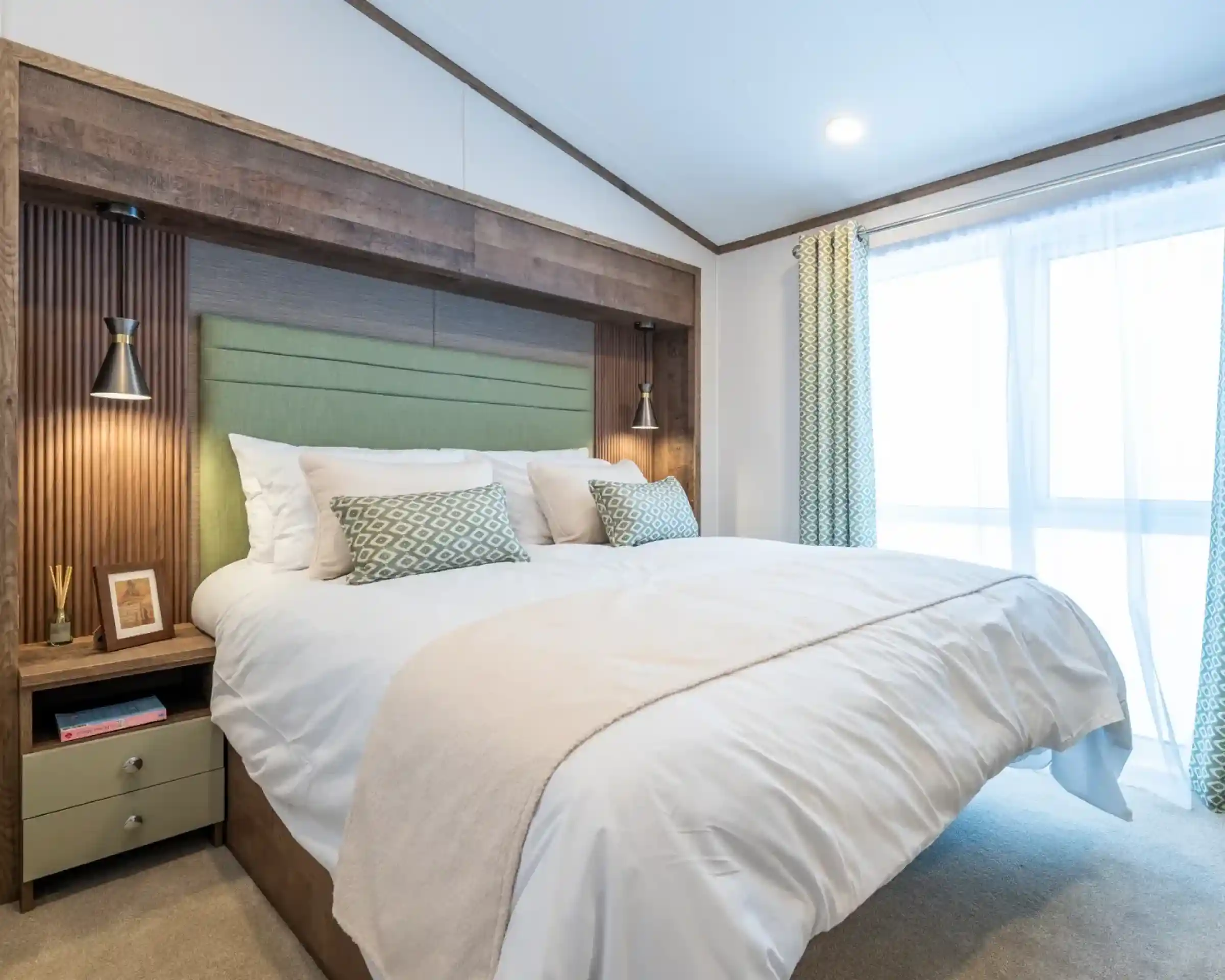 A modern bedroom featuring a king-sized bed with a light green headboard and decorative pillows. On either side of the bed are stylish lamps. The room has a neutral color palette with soft lighting and a large window dressed with sheer curtains. A bedside table with decorative items is visible.