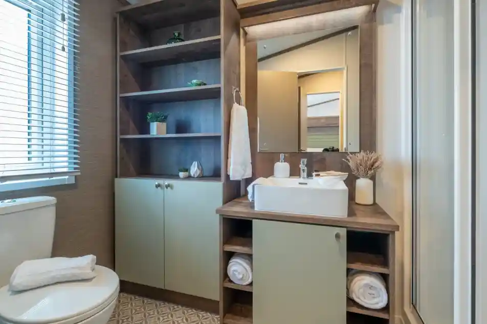 A modern bathroom featuring a white toilet, a wooden vanity with a rectangular sink, and open shelving displaying decorative plants. The walls are light-colored, and there is a mirror above the sink. Towels are neatly arranged on the vanity, and a shower stall is visible in the corner.