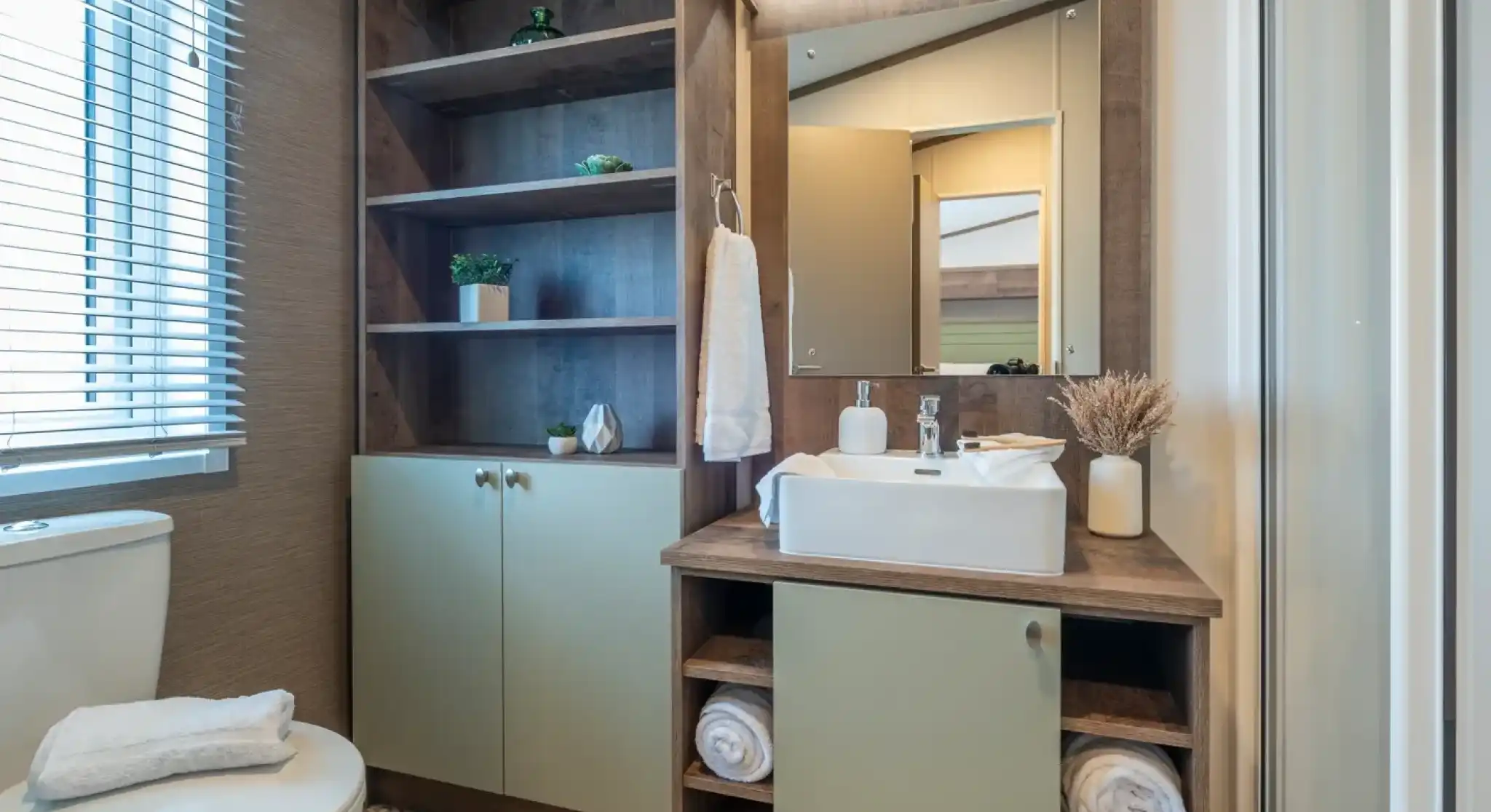 A modern bathroom featuring a white toilet, a wooden vanity with a rectangular sink, and open shelving displaying decorative plants. The walls are light-colored, and there is a mirror above the sink. Towels are neatly arranged on the vanity, and a shower stall is visible in the corner.