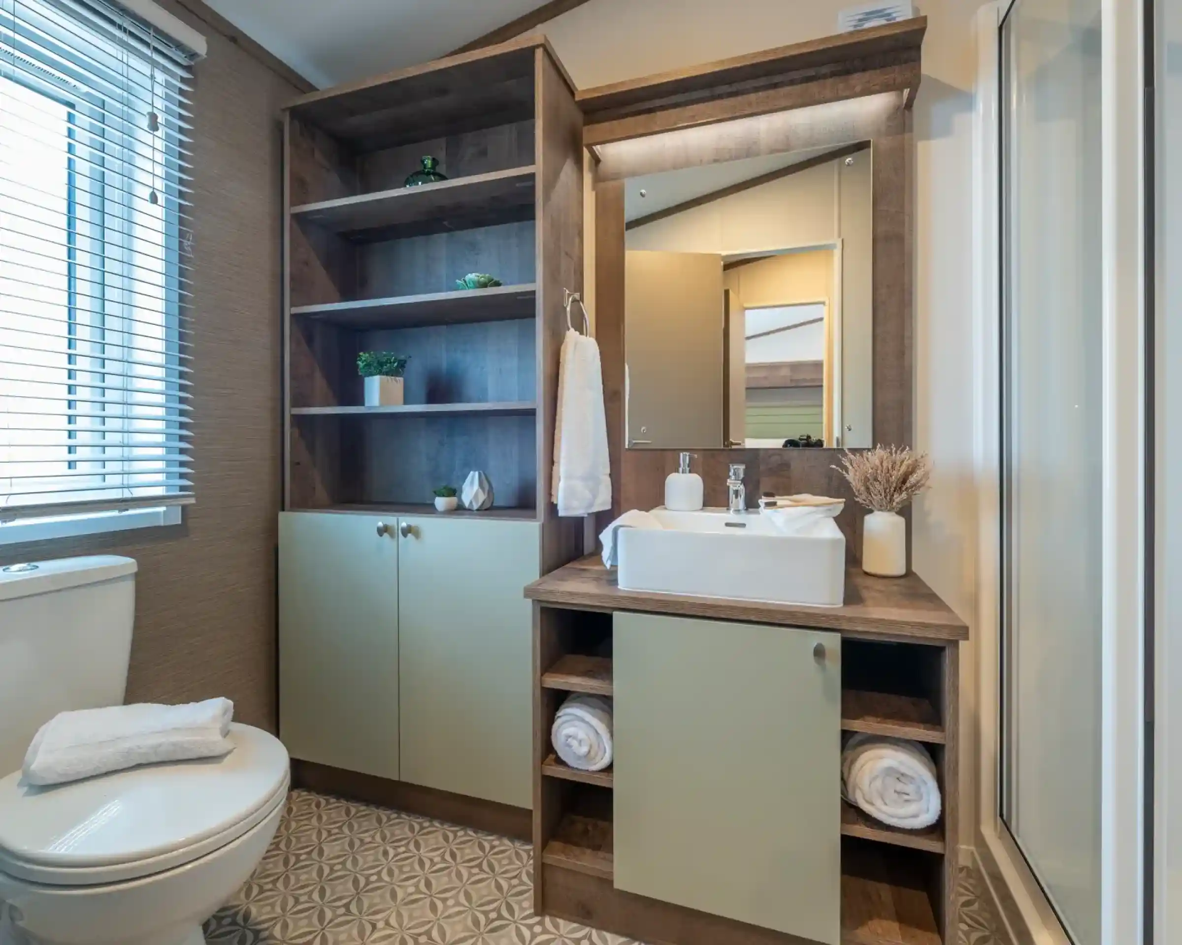 A modern bathroom featuring a white toilet, a wooden vanity with a rectangular sink, and open shelving displaying decorative plants. The walls are light-colored, and there is a mirror above the sink. Towels are neatly arranged on the vanity, and a shower stall is visible in the corner.
