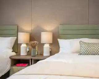 Two twin beds with green upholstered headboards, each accompanied by minimalistic white lamps on wooden nightstands. A small decorative vase with dried flowers and a green bottle sit on the nightstand, adding a touch of warmth to the cozy room.