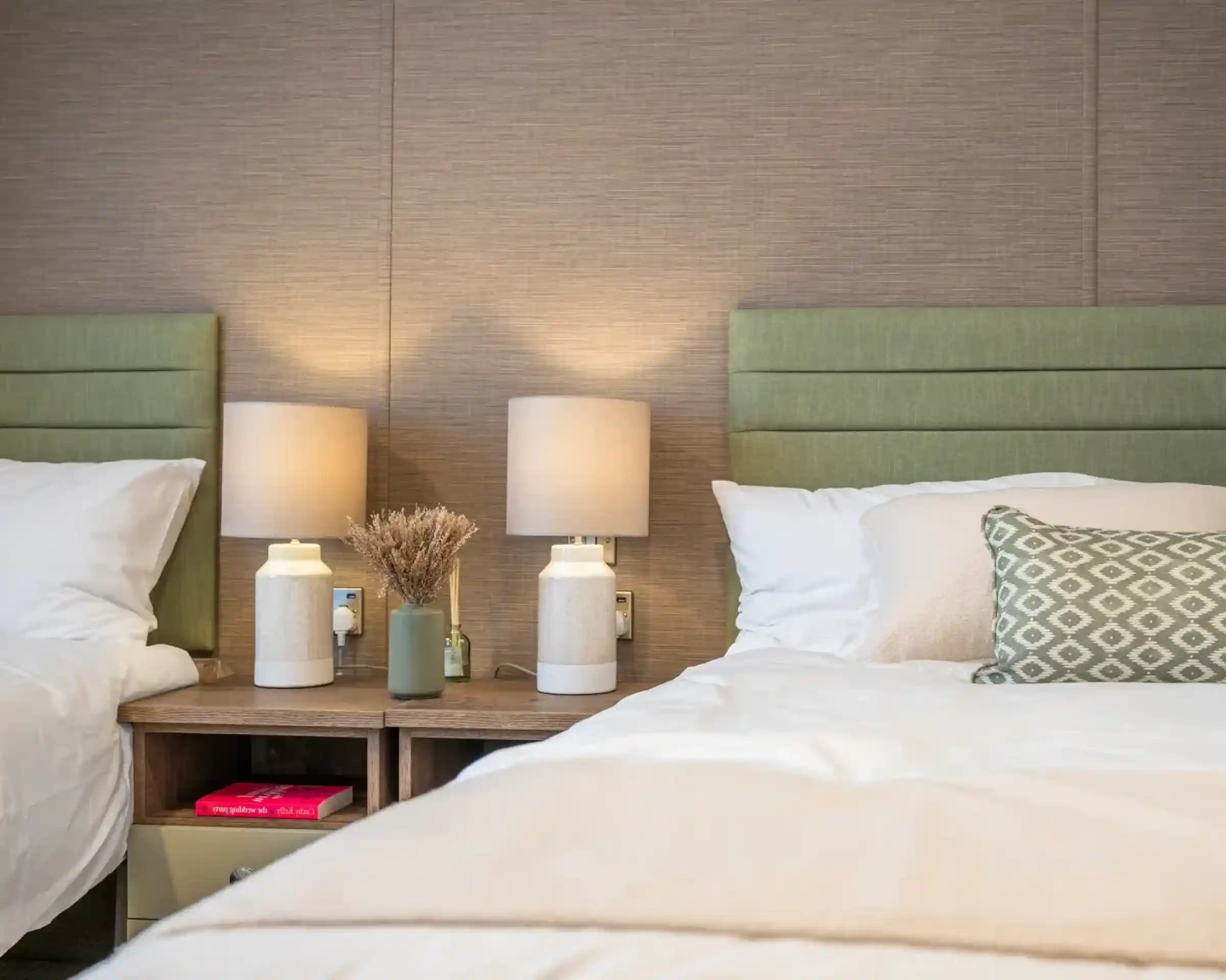 Two twin beds with green upholstered headboards, each accompanied by minimalistic white lamps on wooden nightstands. A small decorative vase with dried flowers and a green bottle sit on the nightstand, adding a touch of warmth to the cozy room.
