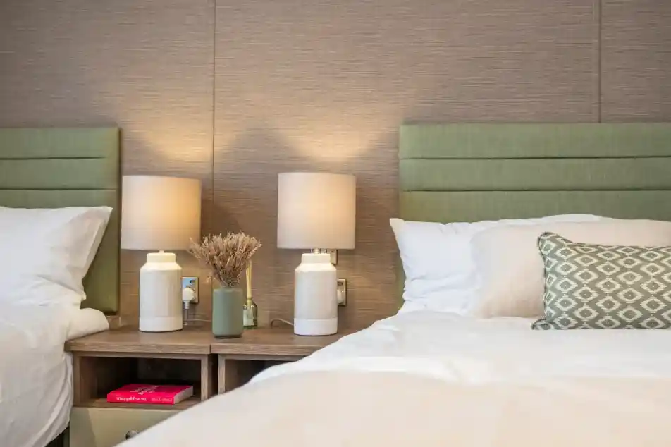 Two twin beds with green upholstered headboards, each accompanied by minimalistic white lamps on wooden nightstands. A small decorative vase with dried flowers and a green bottle sit on the nightstand, adding a touch of warmth to the cozy room.