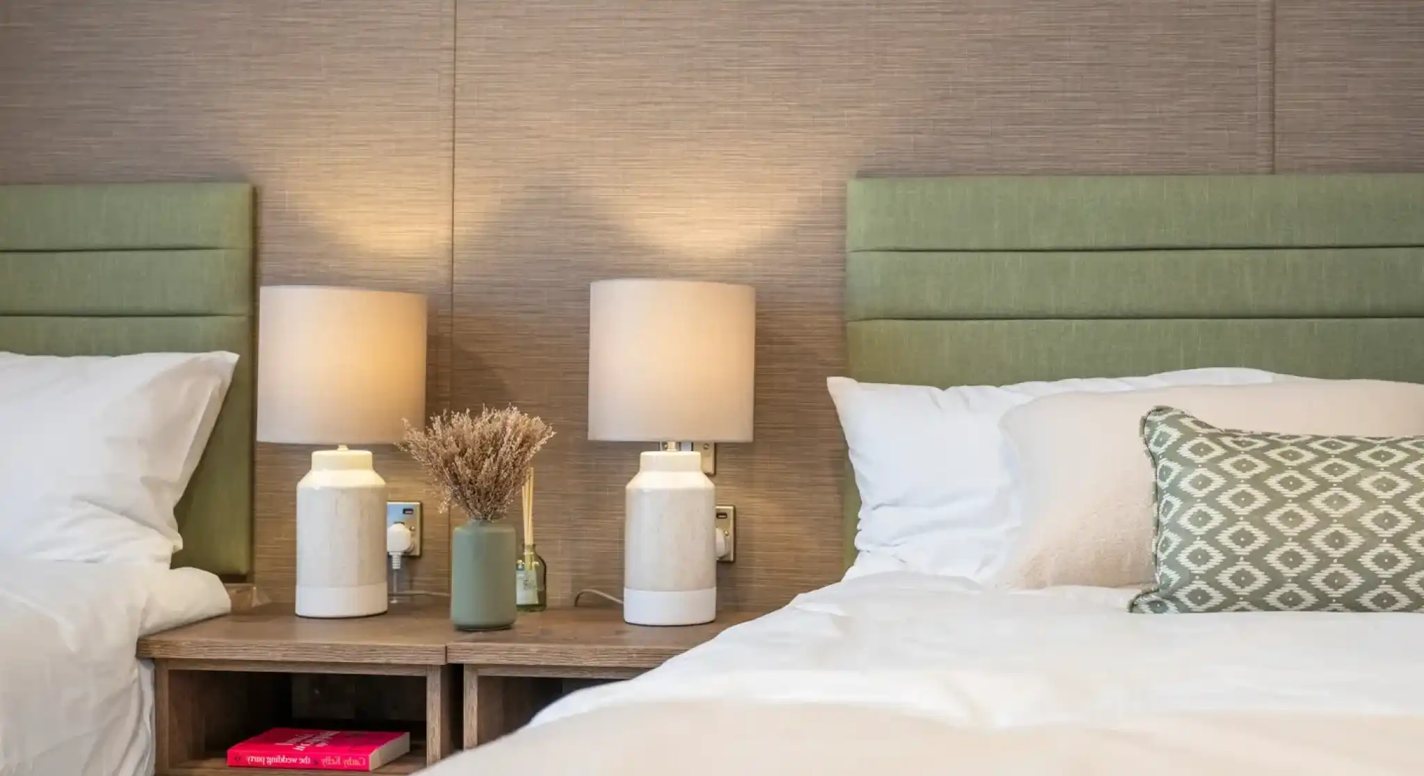 Two twin beds with green upholstered headboards, each accompanied by minimalistic white lamps on wooden nightstands. A small decorative vase with dried flowers and a green bottle sit on the nightstand, adding a touch of warmth to the cozy room.