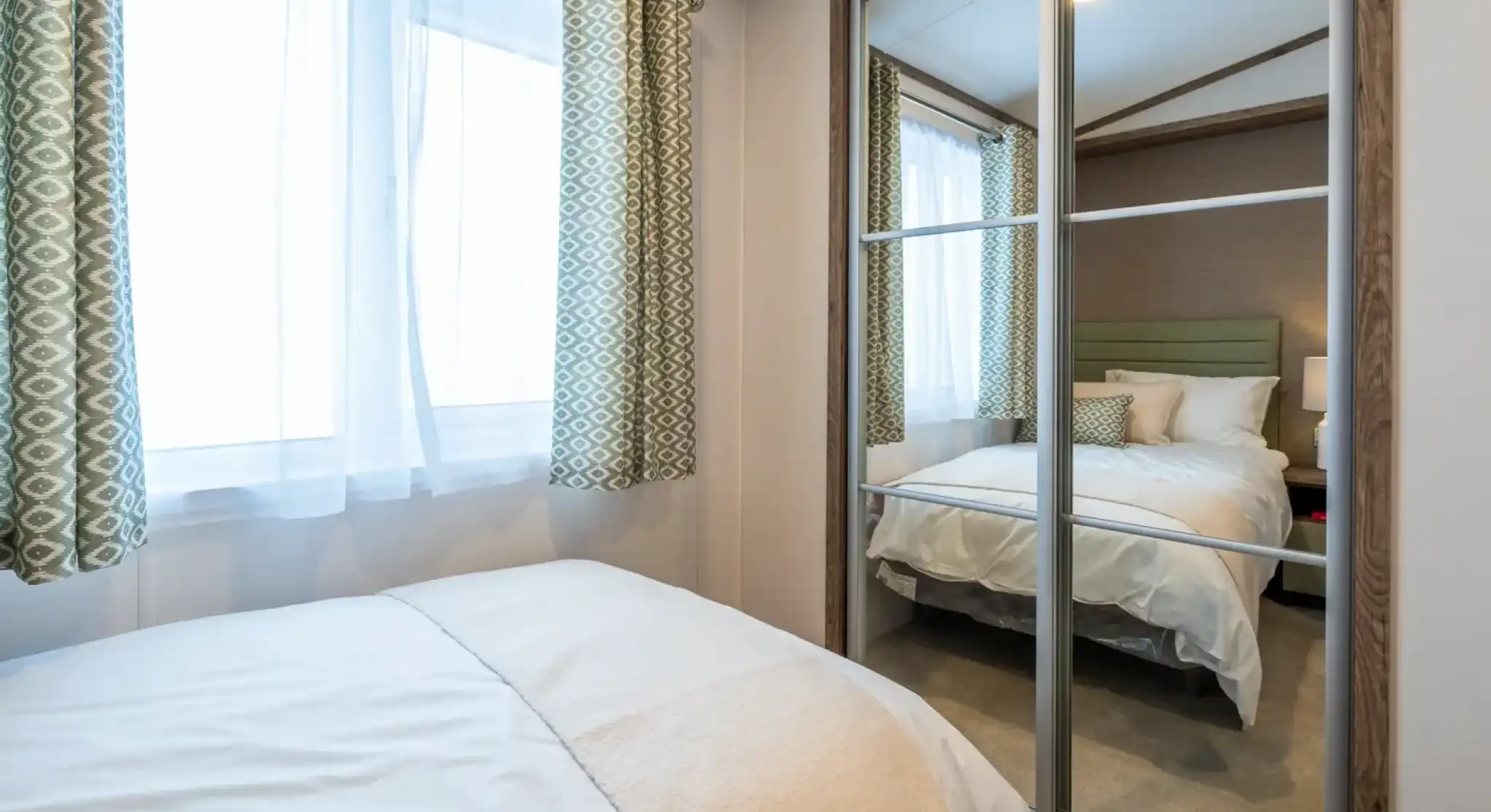 A cozy bedroom featuring a bed with white bedding and a soft throw blanket. Light streams through two large windows with patterned green curtains. A mirrored closet reflects a neatly made bed in the background.