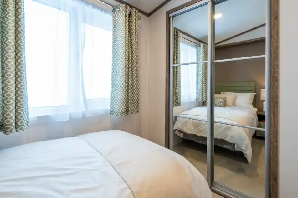 A cozy bedroom featuring a bed with white bedding and a soft throw blanket. Light streams through two large windows with patterned green curtains. A mirrored closet reflects a neatly made bed in the background.