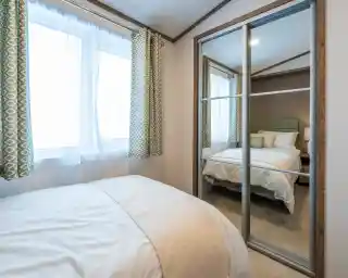 A cozy bedroom featuring a bed with white bedding and a soft throw blanket. Light streams through two large windows with patterned green curtains. A mirrored closet reflects a neatly made bed in the background.