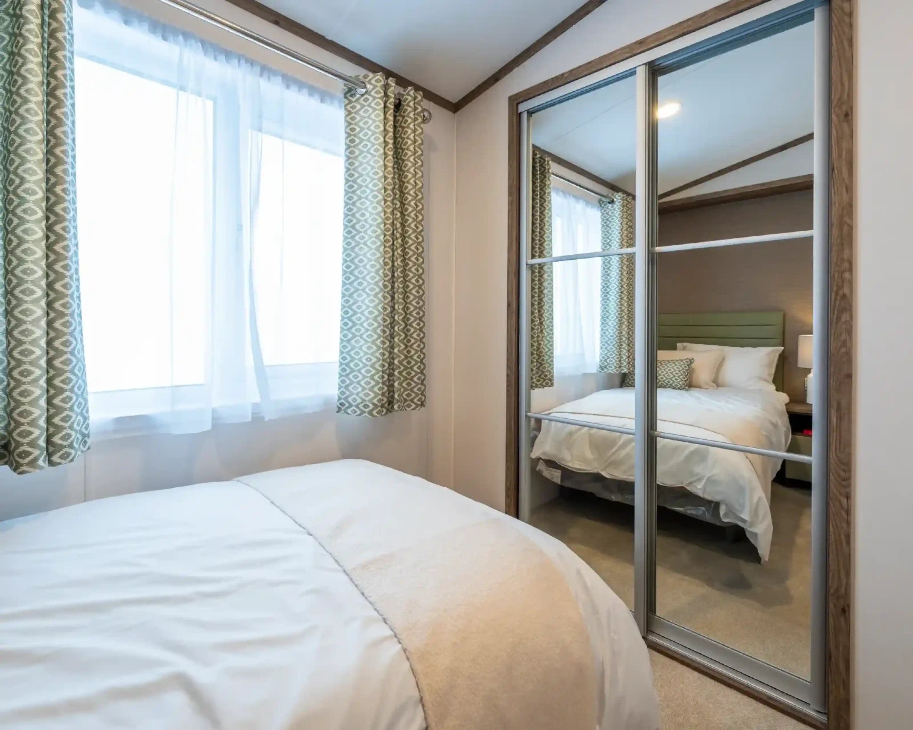 A cozy bedroom featuring a bed with white bedding and a soft throw blanket. Light streams through two large windows with patterned green curtains. A mirrored closet reflects a neatly made bed in the background.