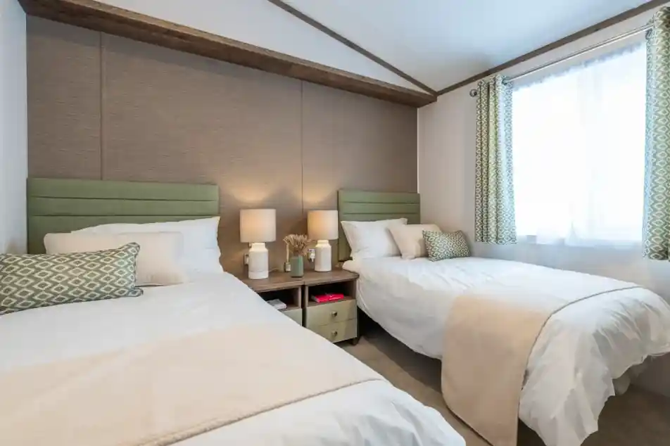 A cozy bedroom featuring two twin beds with light green headboards, white linens, and decorative pillows. A small nightstand between the beds holds two lamps. A window with sheer curtains lets in natural light, and there’s a beige throw blanket draped over one bed. The room has a neutral color palette and minimal decor.