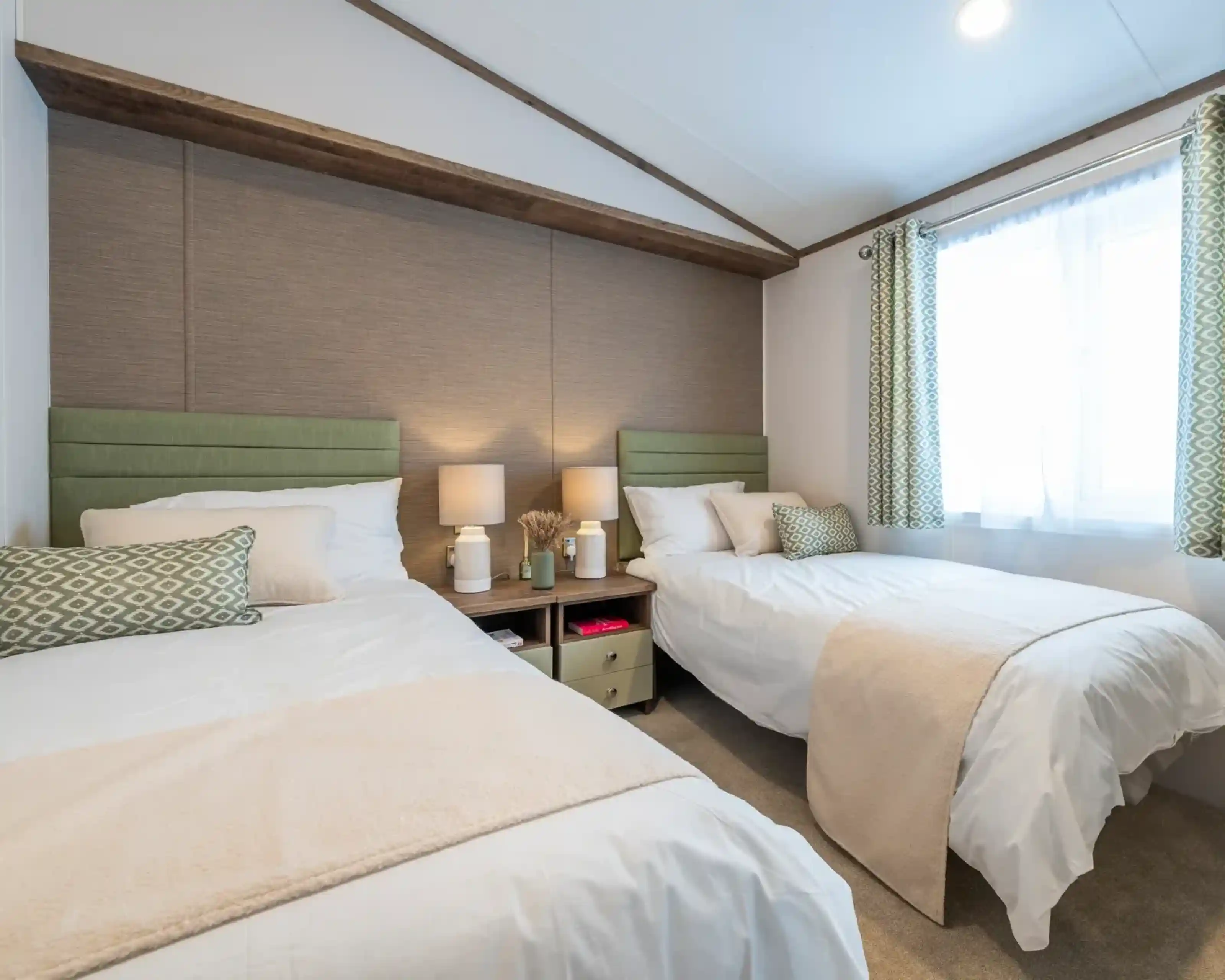 A cozy bedroom featuring two twin beds with light green headboards, white linens, and decorative pillows. A small nightstand between the beds holds two lamps. A window with sheer curtains lets in natural light, and there’s a beige throw blanket draped over one bed. The room has a neutral color palette and minimal decor.