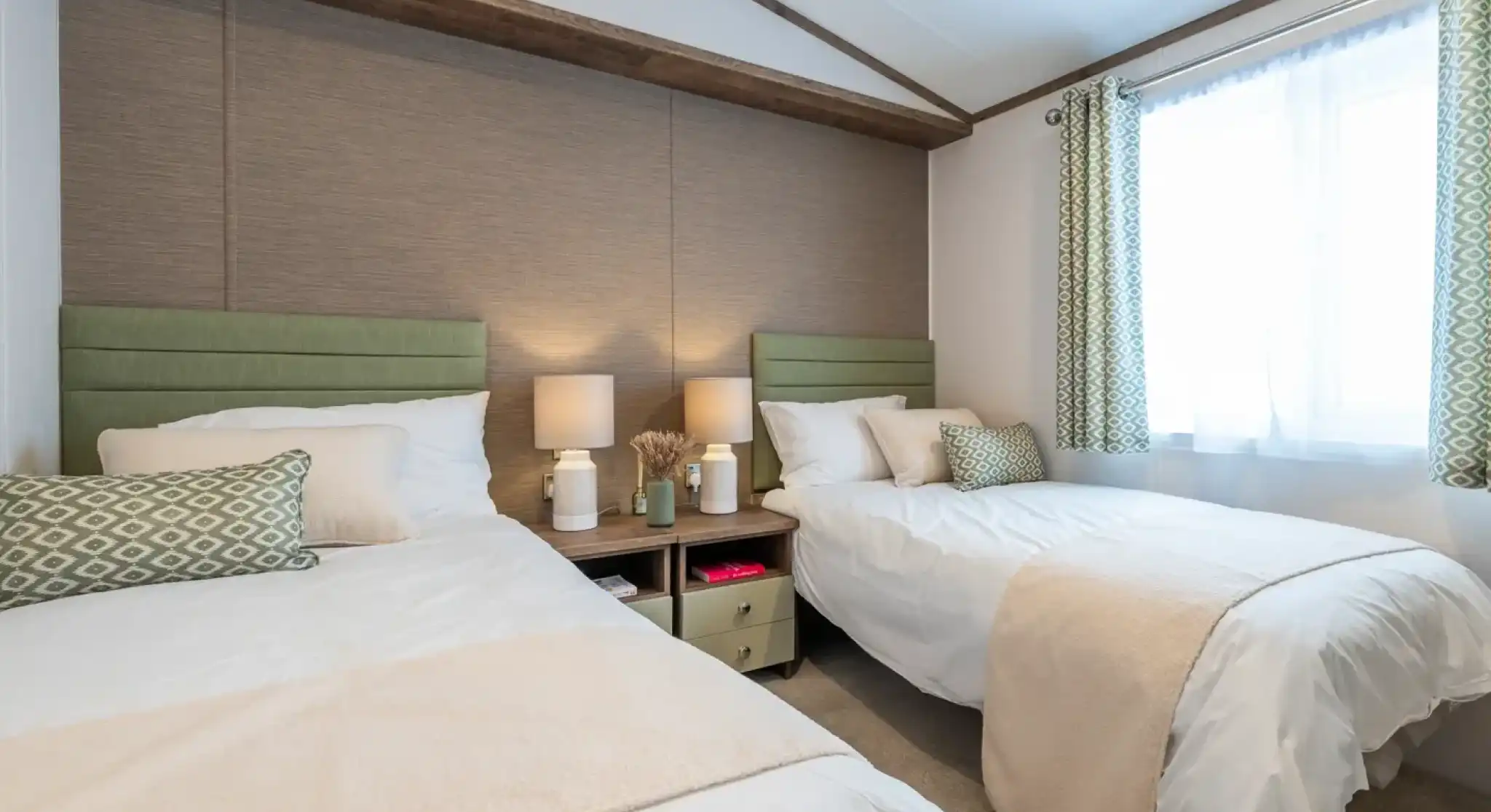 A cozy bedroom featuring two twin beds with light green headboards, white linens, and decorative pillows. A small nightstand between the beds holds two lamps. A window with sheer curtains lets in natural light, and there’s a beige throw blanket draped over one bed. The room has a neutral color palette and minimal decor.