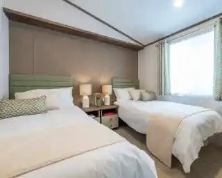 A cozy bedroom featuring two twin beds with light green headboards, white linens, and decorative pillows. A small nightstand between the beds holds two lamps. A window with sheer curtains lets in natural light, and there’s a beige throw blanket draped over one bed. The room has a neutral color palette and minimal decor.
