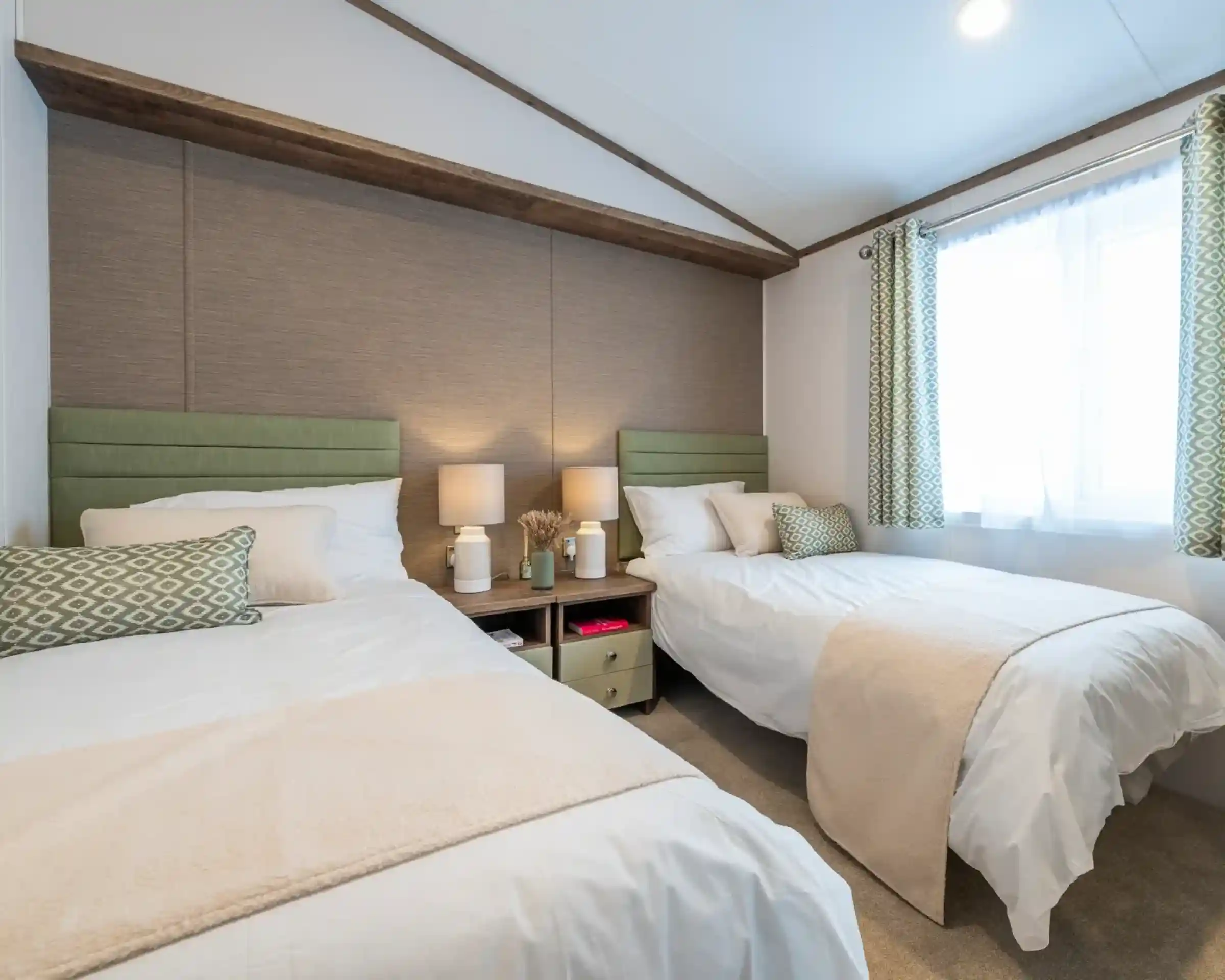 A cozy bedroom featuring two twin beds with light green headboards, white linens, and decorative pillows. A small nightstand between the beds holds two lamps. A window with sheer curtains lets in natural light, and there’s a beige throw blanket draped over one bed. The room has a neutral color palette and minimal decor.