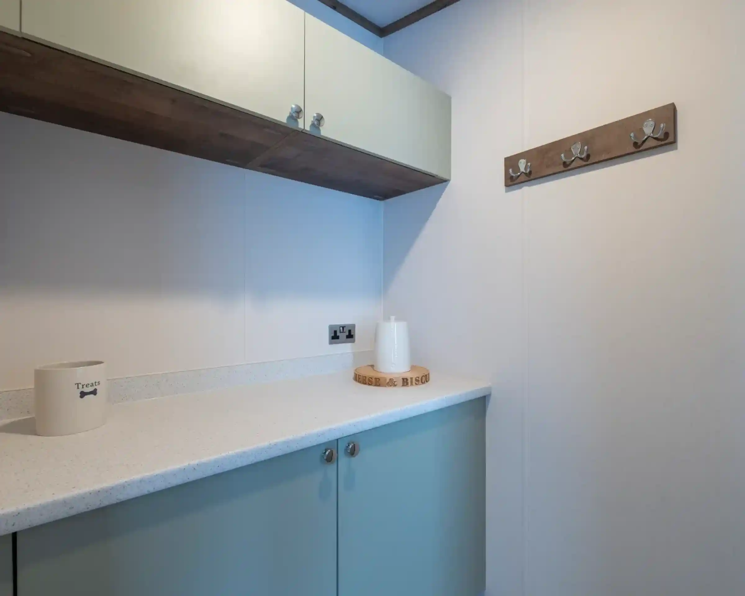 A modern, minimalist laundry area featuring light-colored cabinetry, a white countertop, and a decorative wooden coat rack with hooks on the wall. A small round wooden tray and a decorative white object are placed on the counter.