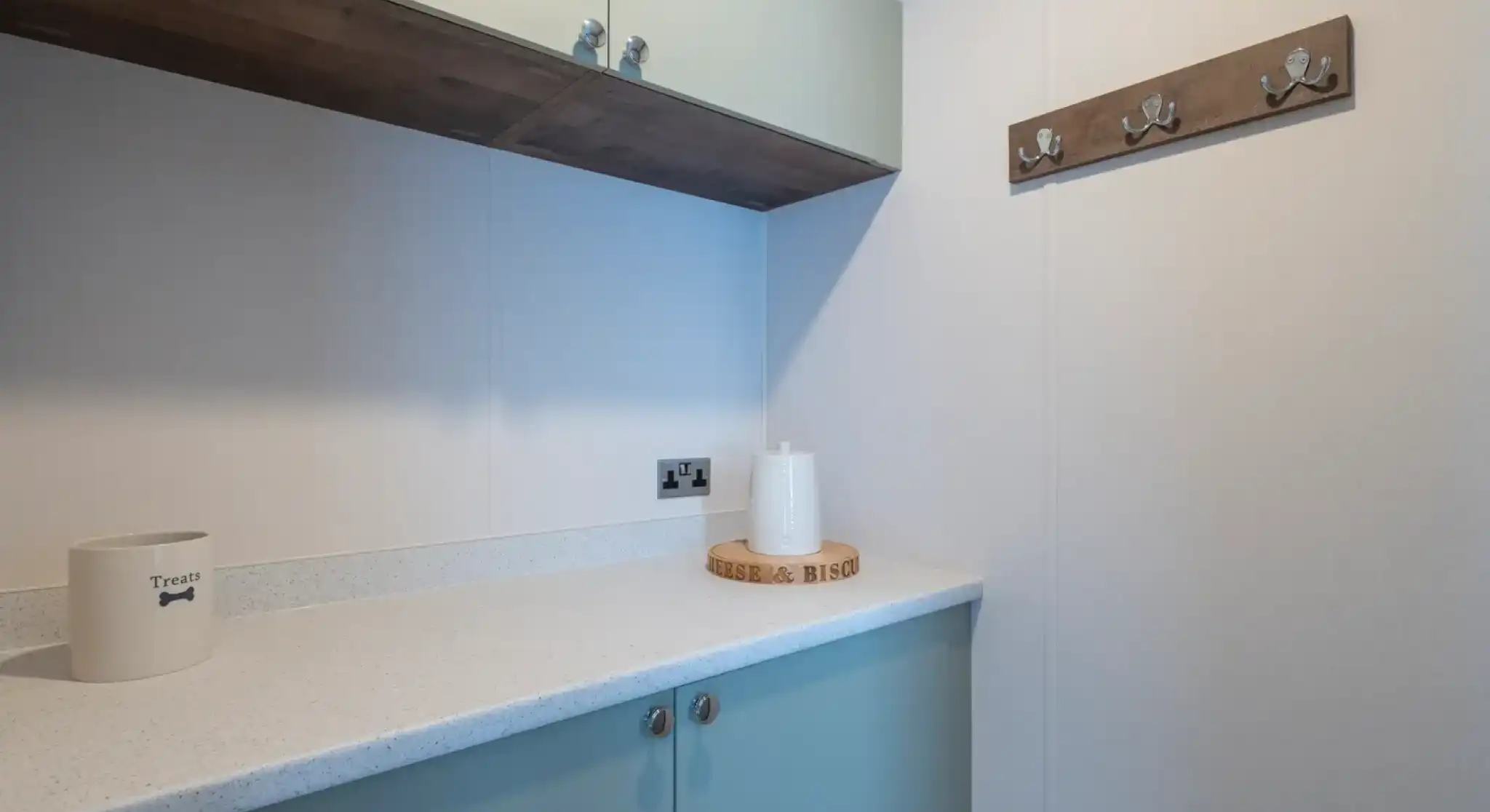 A modern, minimalist laundry area featuring light-colored cabinetry, a white countertop, and a decorative wooden coat rack with hooks on the wall. A small round wooden tray and a decorative white object are placed on the counter.