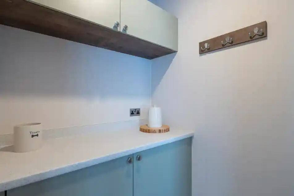 A modern, minimalist laundry area featuring light-colored cabinetry, a white countertop, and a decorative wooden coat rack with hooks on the wall. A small round wooden tray and a decorative white object are placed on the counter.