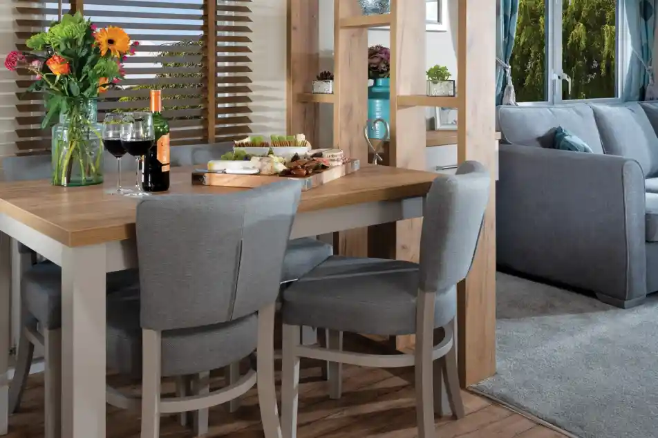 Modern dining area with a wooden table, grey chairs, and colourful flowers. Cheese and wine set a cosy, inviting mood. Bright window view outside.