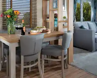 Modern dining area with a wooden table, grey chairs, and colourful flowers. Cheese and wine set a cosy, inviting mood. Bright window view outside.