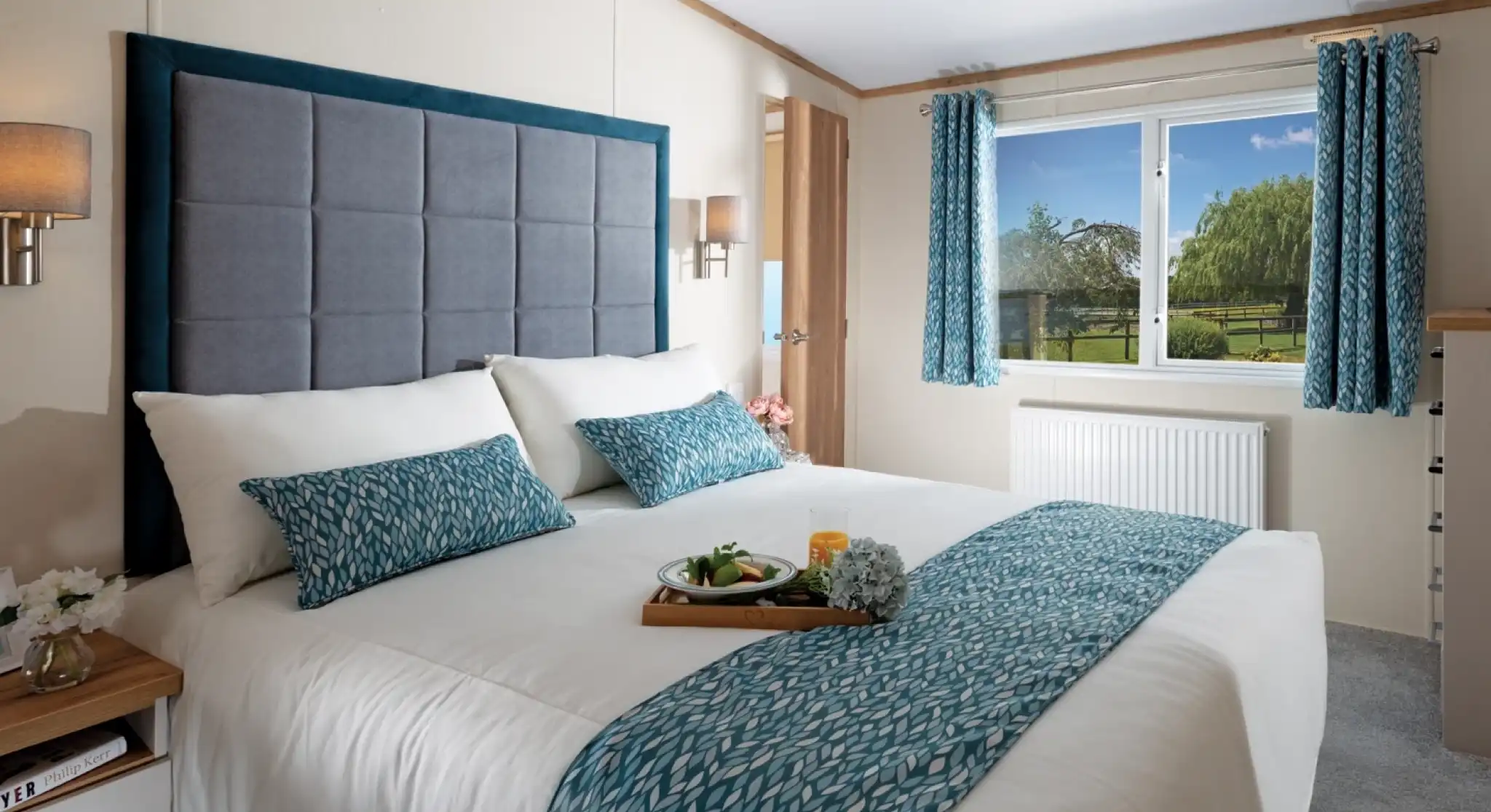 Bright bedroom with a large, neatly made bed featuring blue-patterned accents. A window with matching curtains reveals a scenic garden view. Cosy and inviting atmosphere.