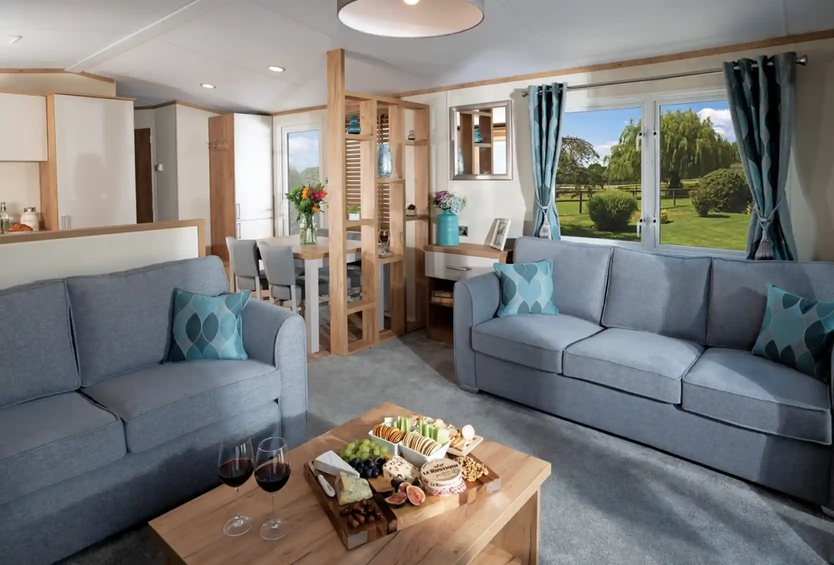 Modern living room with two blue sofas, wood coffee table with snacks and wine; large window showing a lush garden. Bright and inviting atmosphere.