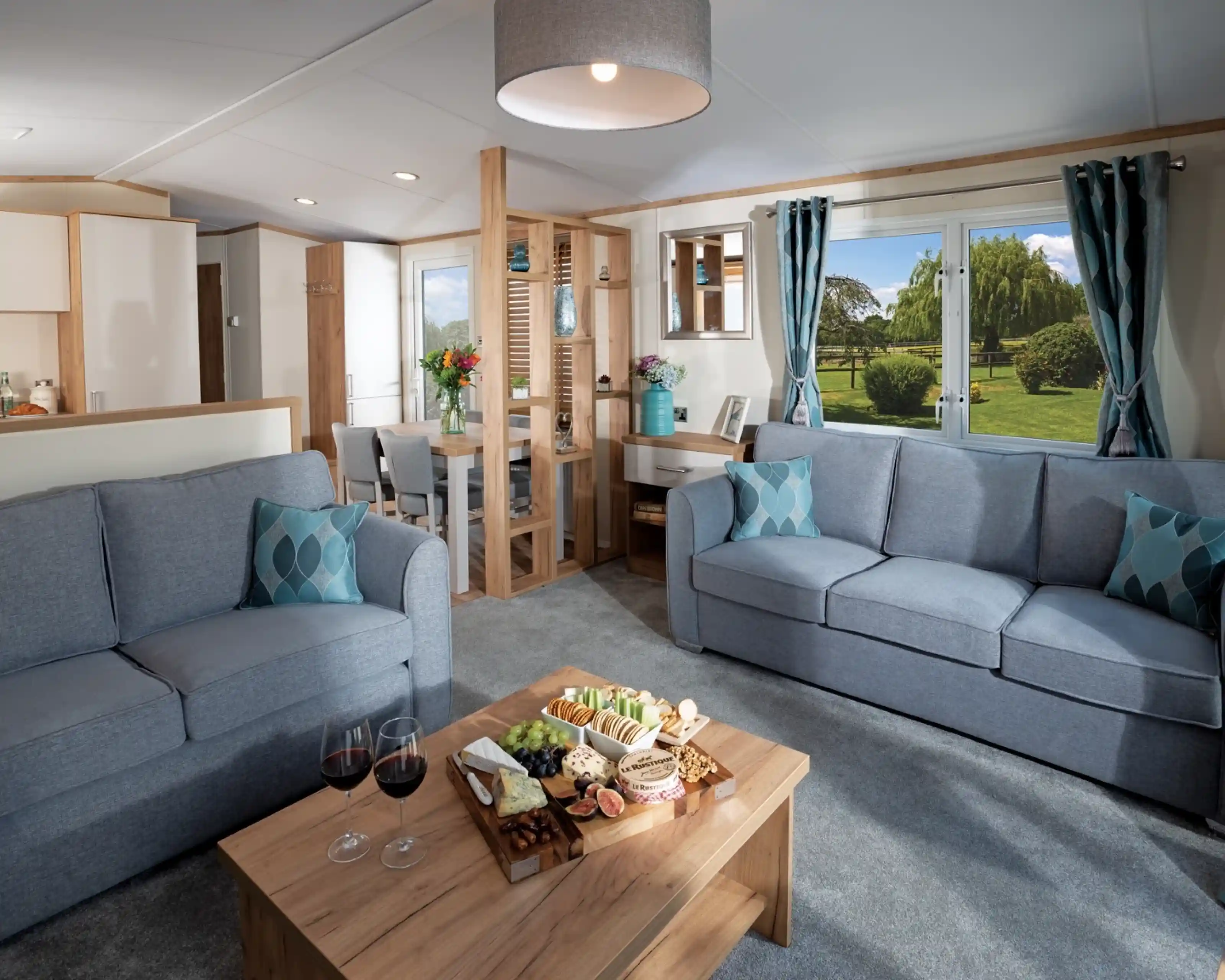 Modern living room with two blue sofas, wood coffee table with snacks and wine; large window showing a lush garden. Bright and inviting atmosphere.