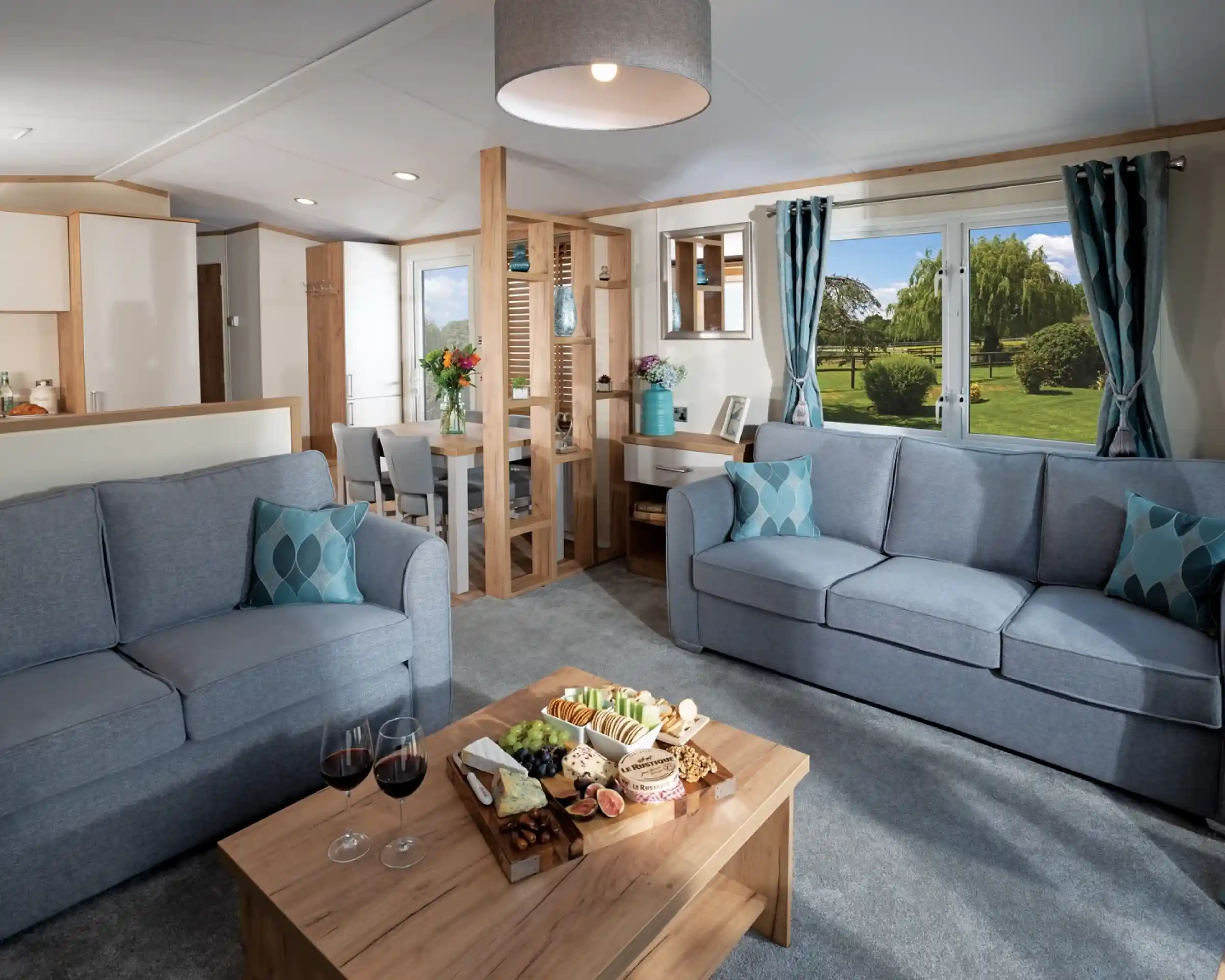 Modern living room with two blue sofas, wood coffee table with snacks and wine; large window showing a lush garden. Bright and inviting atmosphere.