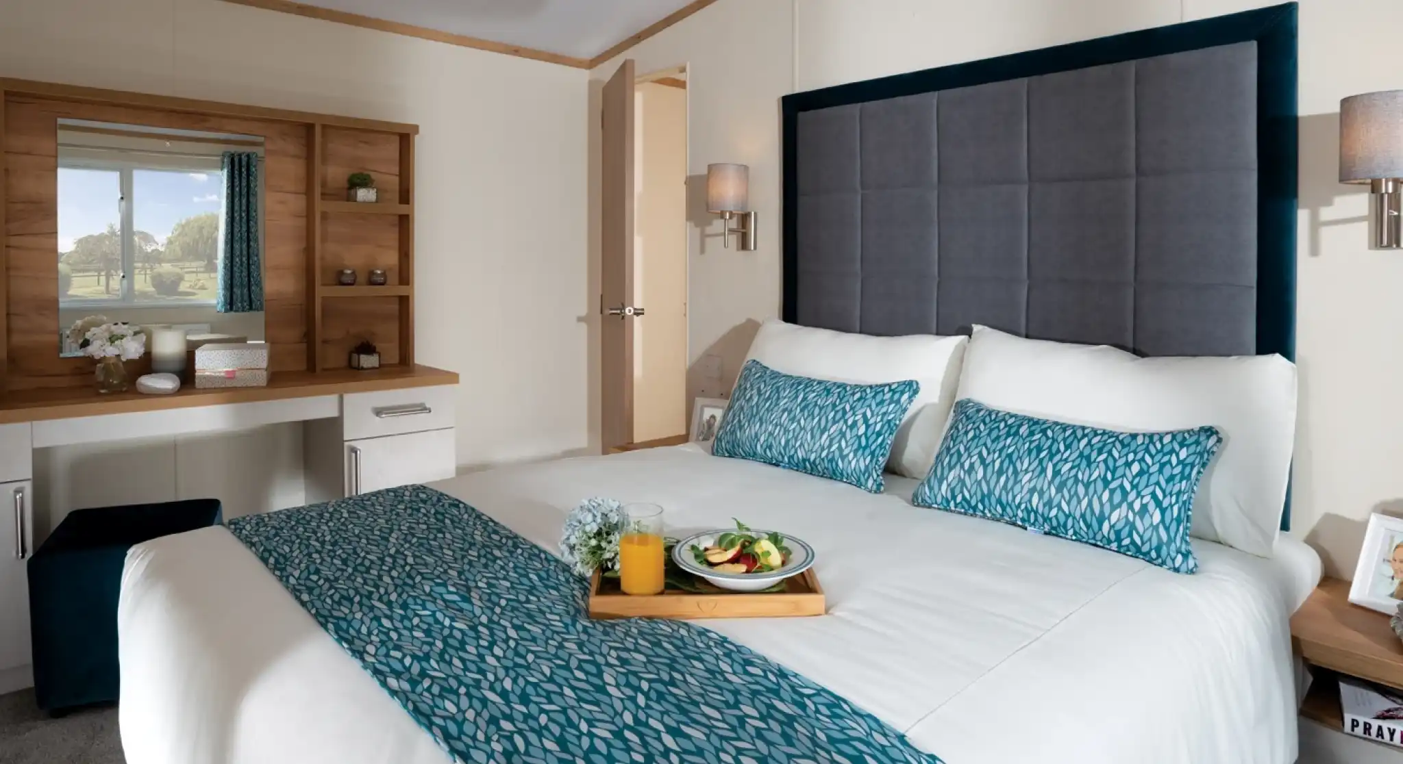 Modern bedroom with a large bed featuring blue patterned accents. A tray with breakfast sits on the bed. A vanity and scenic window view create a serene ambience.