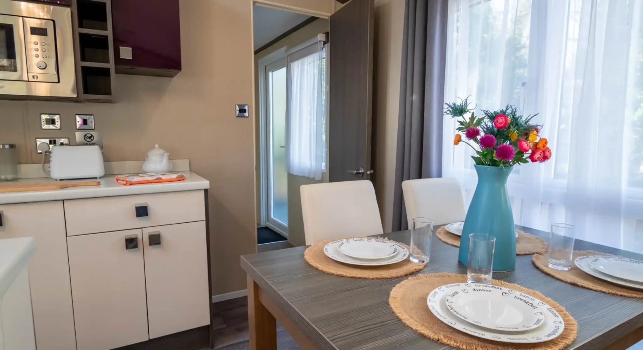 A dining table set for four with four dining chairs, sat in a kitchen with gloss cabinets and a window to the side with curtains.