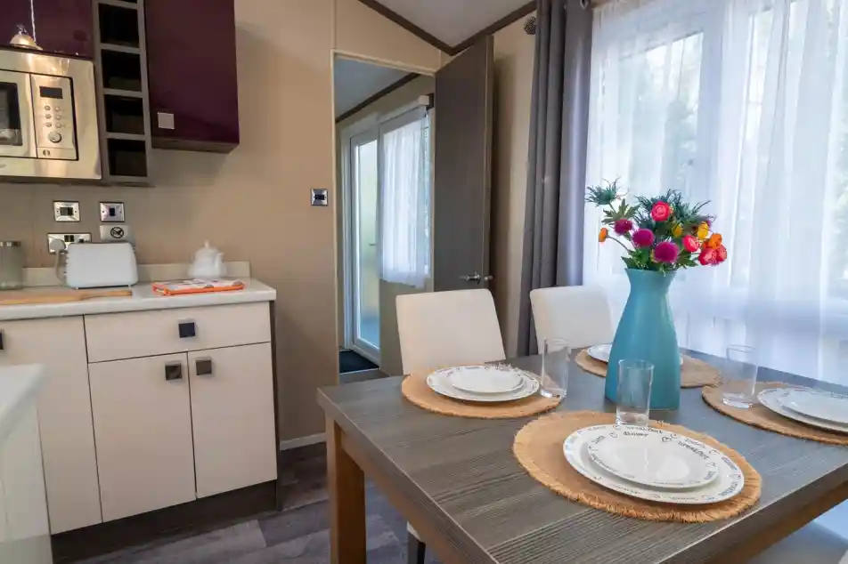 A dining table set for four with four dining chairs, sat in a kitchen with gloss cabinets and a window to the side with curtains.