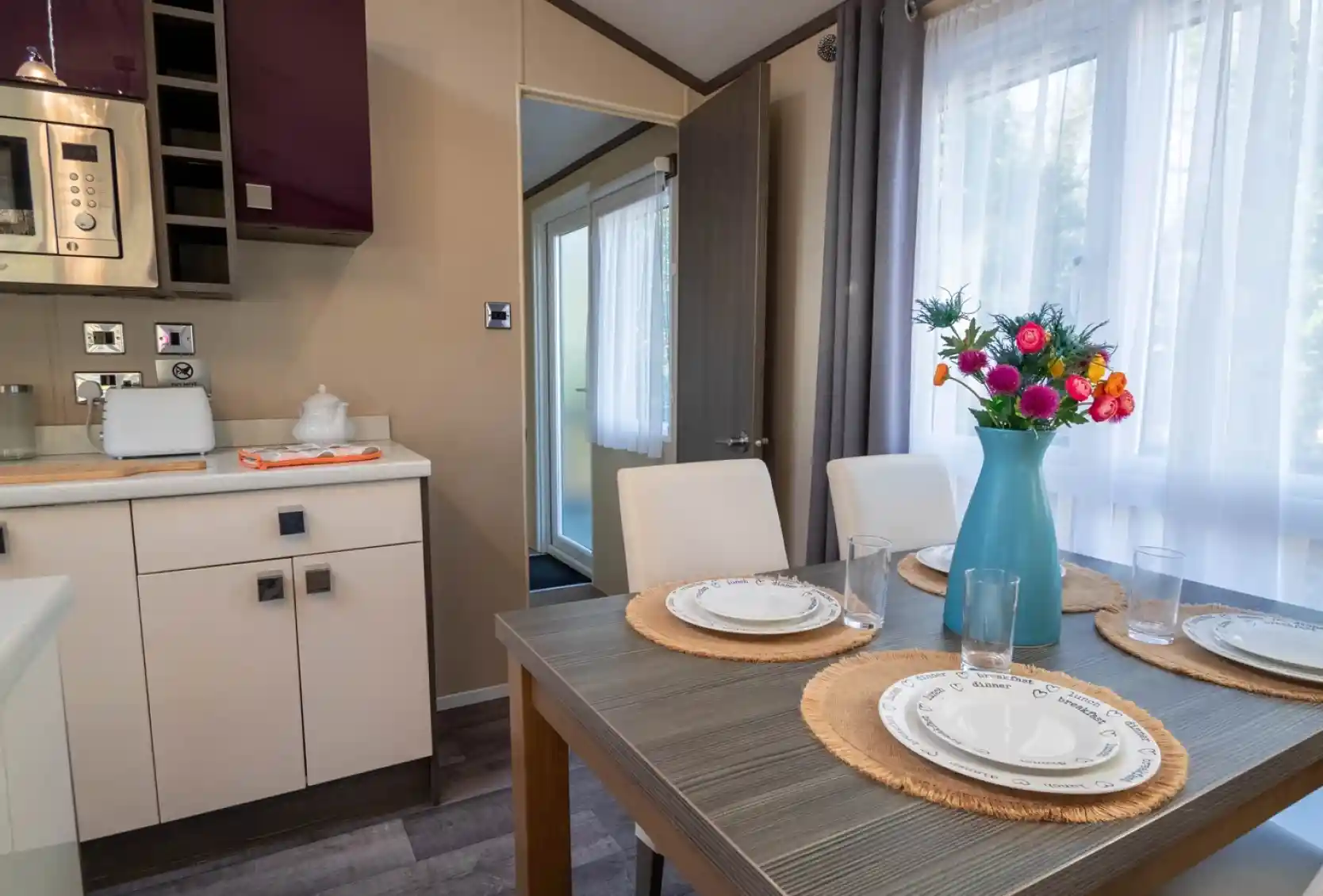 A dining table set for four with four dining chairs, sat in a kitchen with gloss cabinets and a window to the side with curtains.