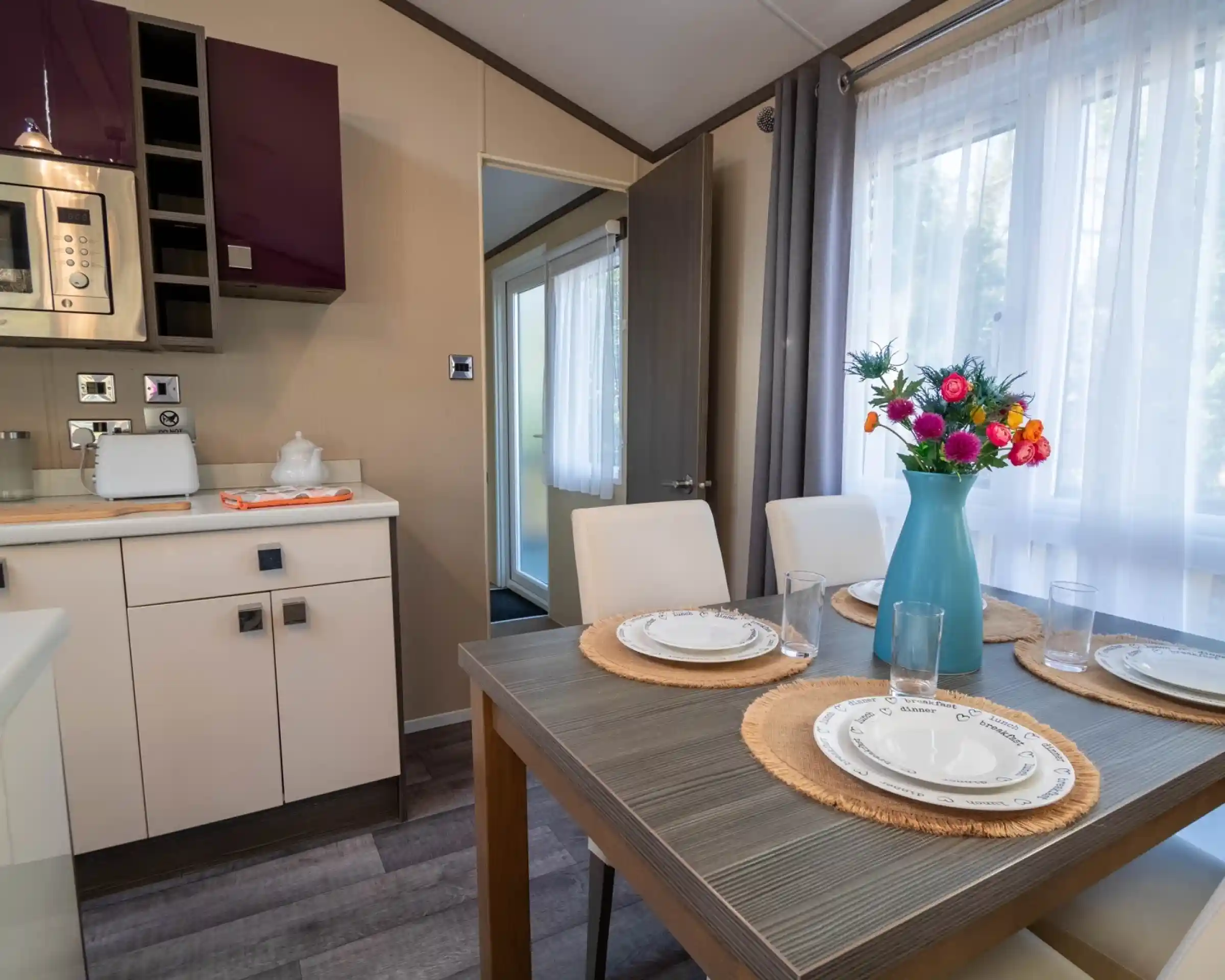 A dining table set for four with four dining chairs, sat in a kitchen with gloss cabinets and a window to the side with curtains.