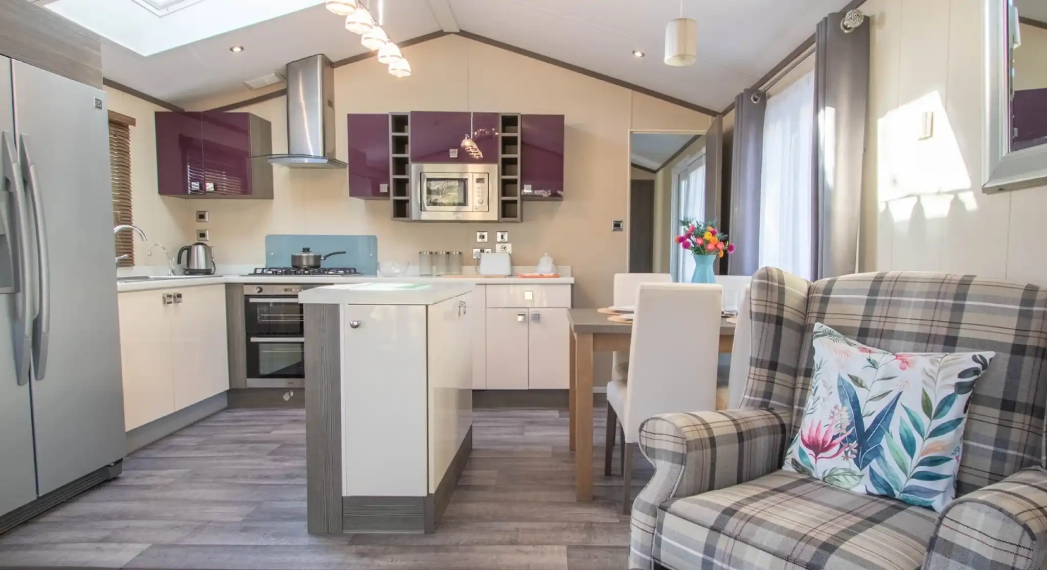 An open plan kitchen, dining, living room with Velux roof window and laminate wood flooring to the kitchen area. A mixture of white gloss and purple gloss cabinets, a kitchen island and a dining table with chairs separates the kitchen area from the lounge.